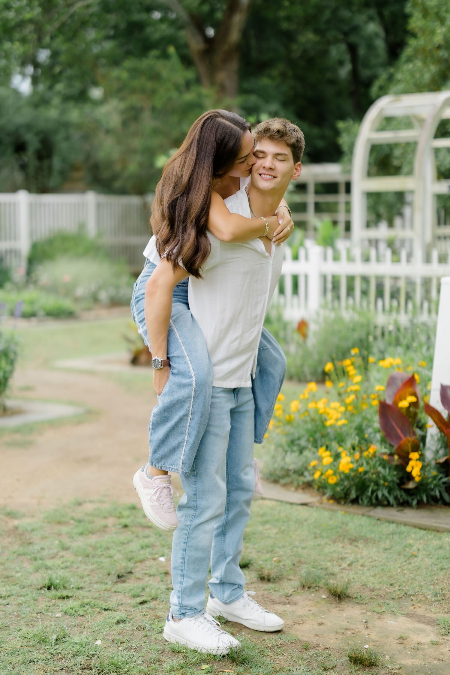 A young couple shares a romantic piggyback ride in a lush garden with yellow flowers and white fence.