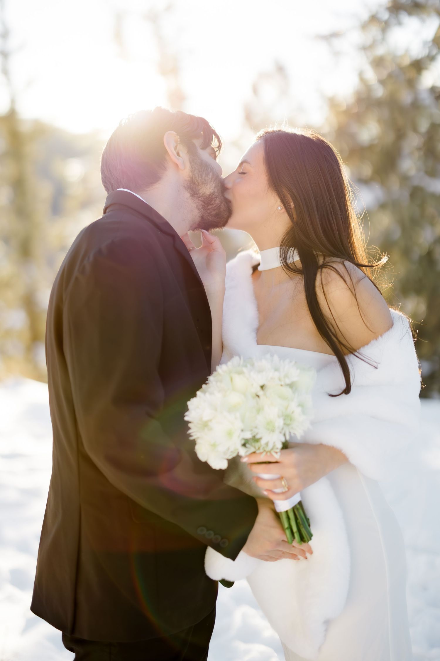 Bride and groom share a kiss in snowy winter landscape, bride holding white bouquet with sun flare behind them.