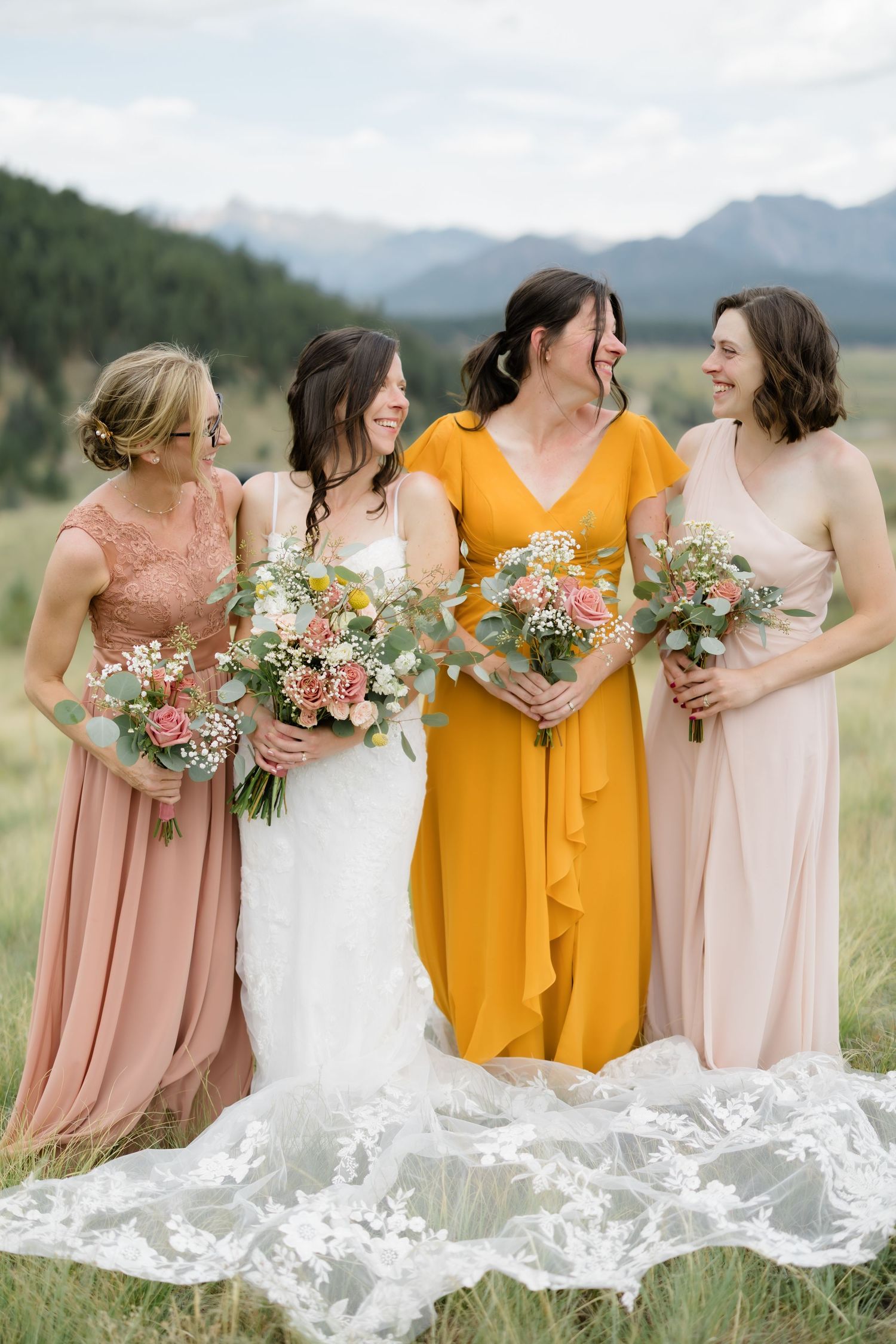 Bride in white lace gown with three bridesmaids in mismatched pink and yellow dresses holding floral bouquets in a mountain meadow.