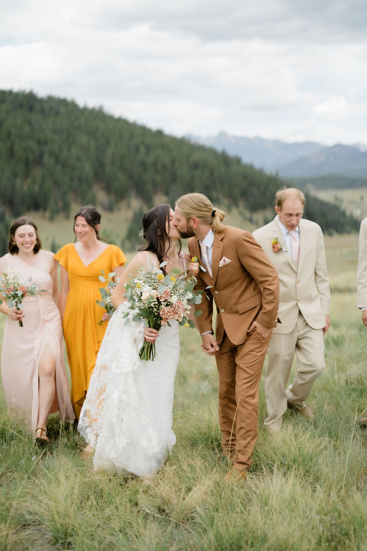 Bride and groom share a kiss while walking with wedding party in a mountain meadow, bridesmaids in yellow and pink gowns.