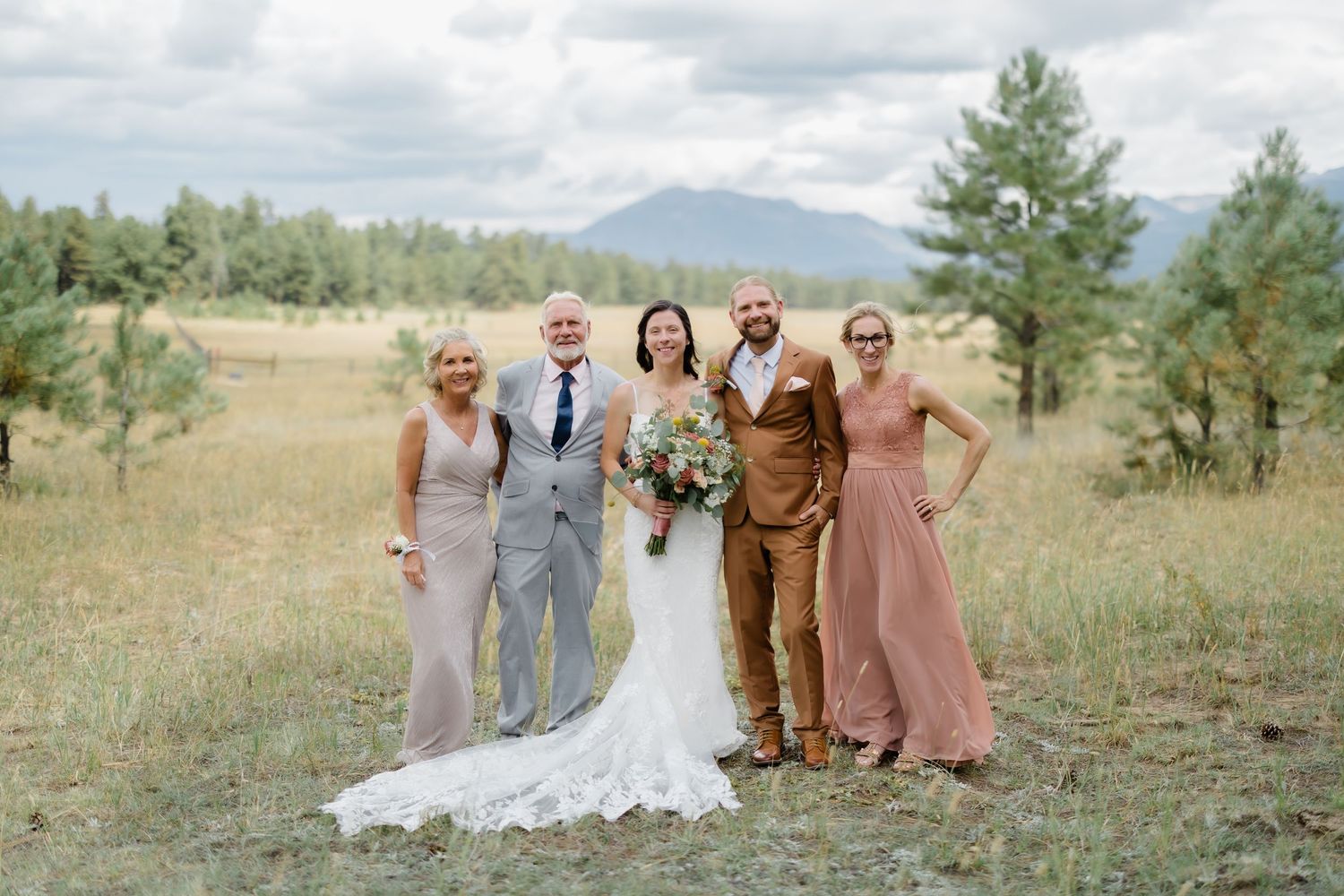 Wedding party posing outdoors in a meadow with mountain backdrop, bride in white lace gown with bouquet, guests in earth tones.