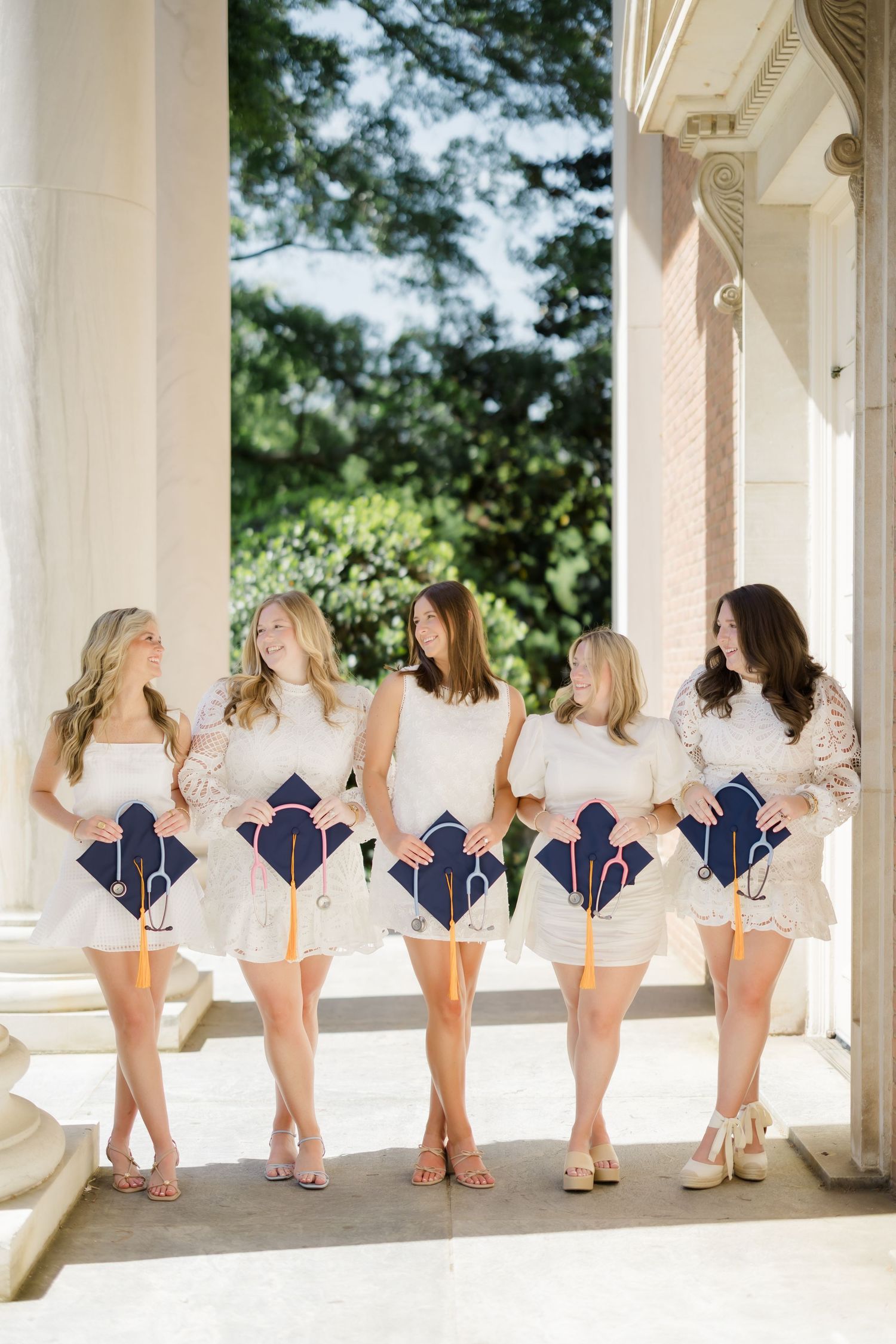Five women in white dresses hold navy graduation caps with gold tassels between classical columns outdoors.
