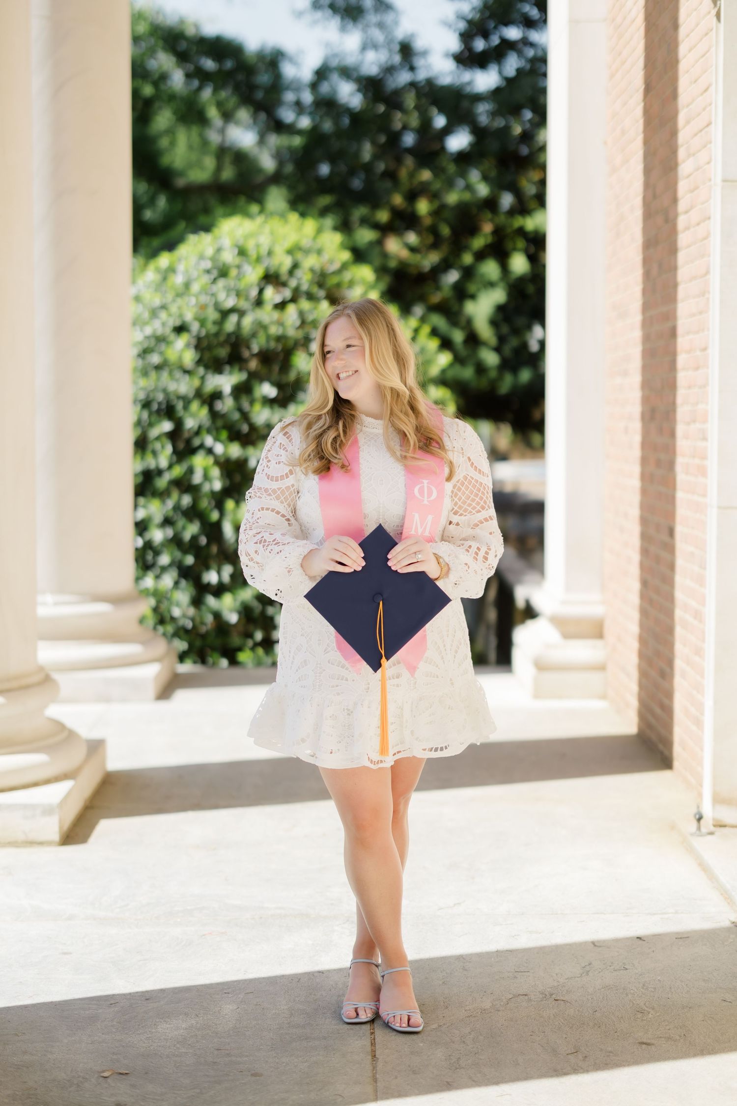 Smiling female graduate in white lace dress holding navy cap with Phi Mu sorority sash between elegant columns.