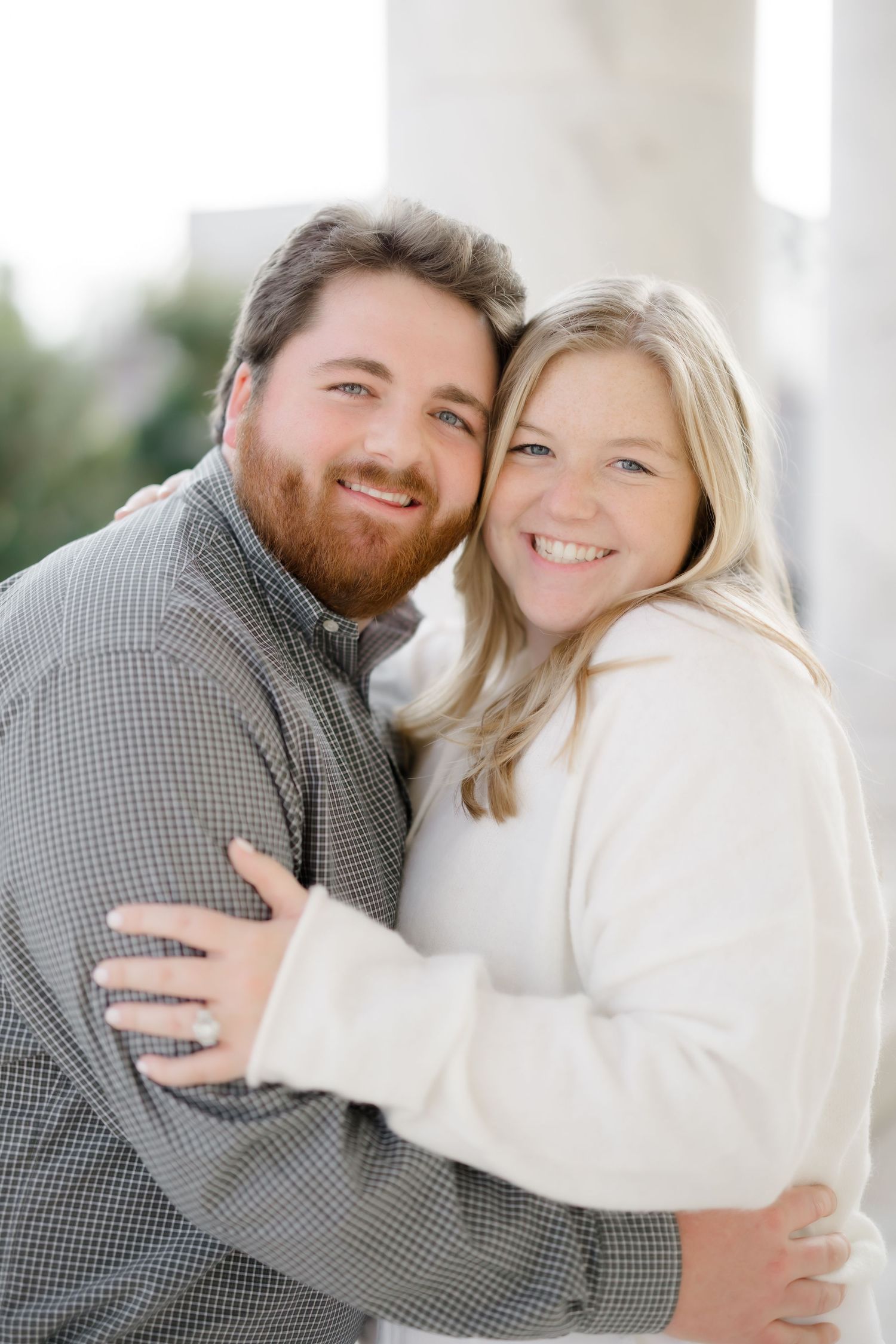 A smiling couple embracing outdoors, man with red beard in gray shirt, woman in white sweater, engagement photo.