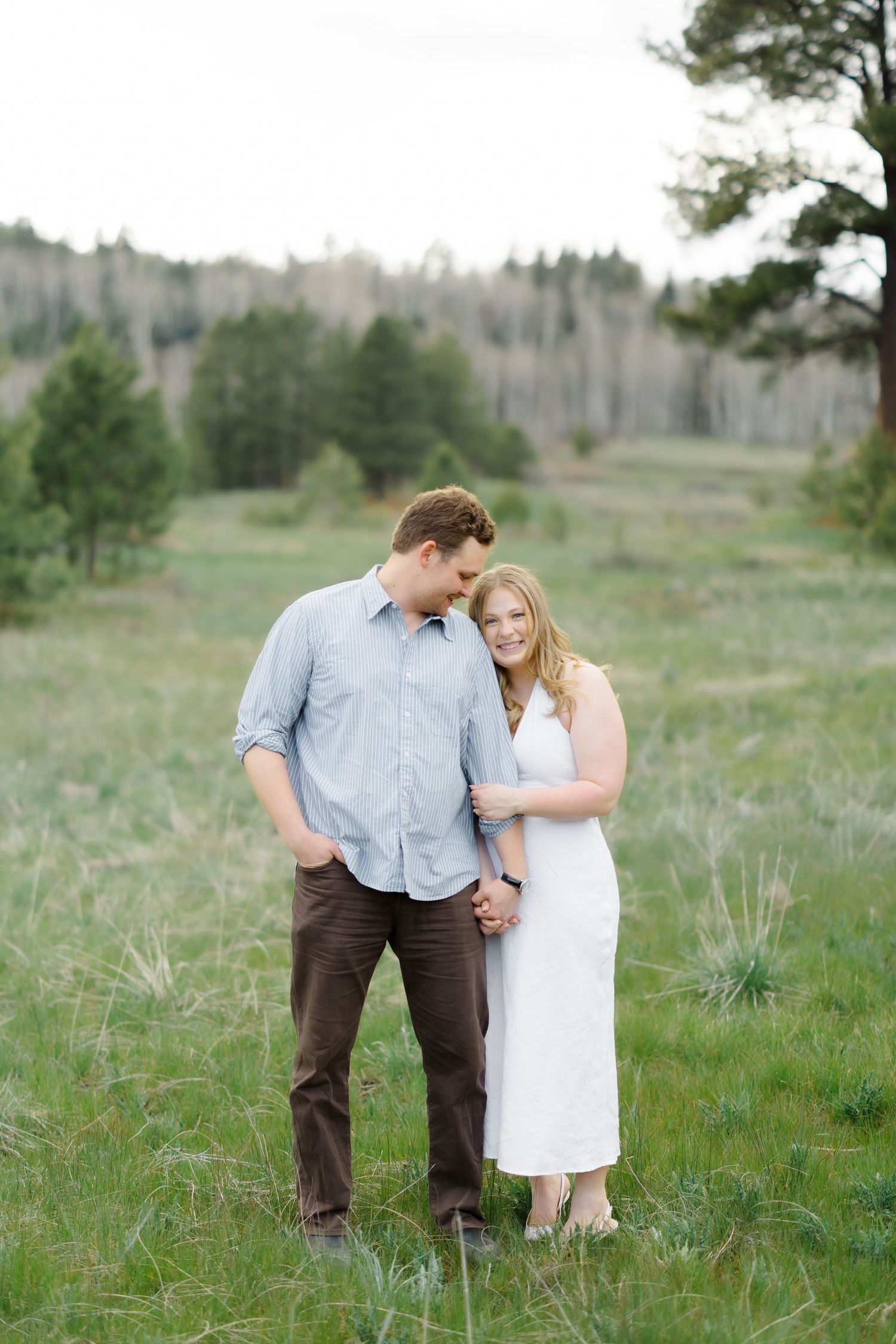 Couple standing close together in a green meadow with pine trees and rolling hills in the background.