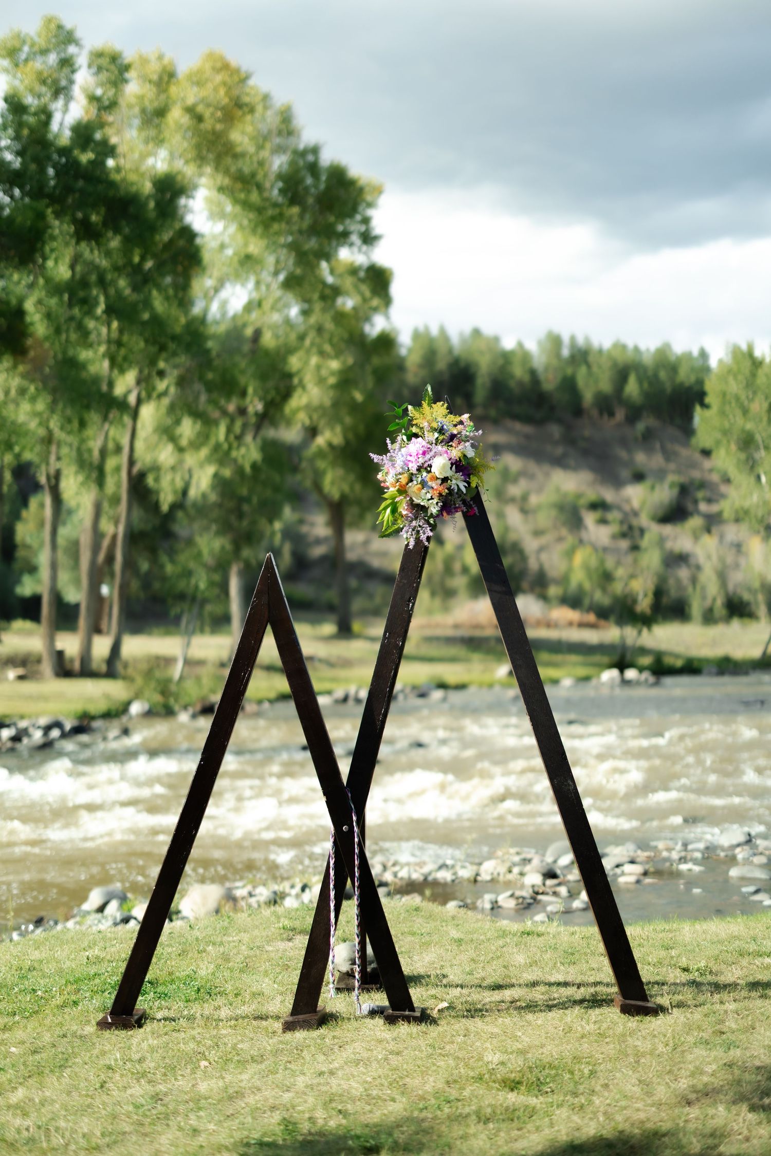 Rustic wooden triangular wedding arch adorned with colorful flowers, set outdoors by a flowing river with lush green trees.
