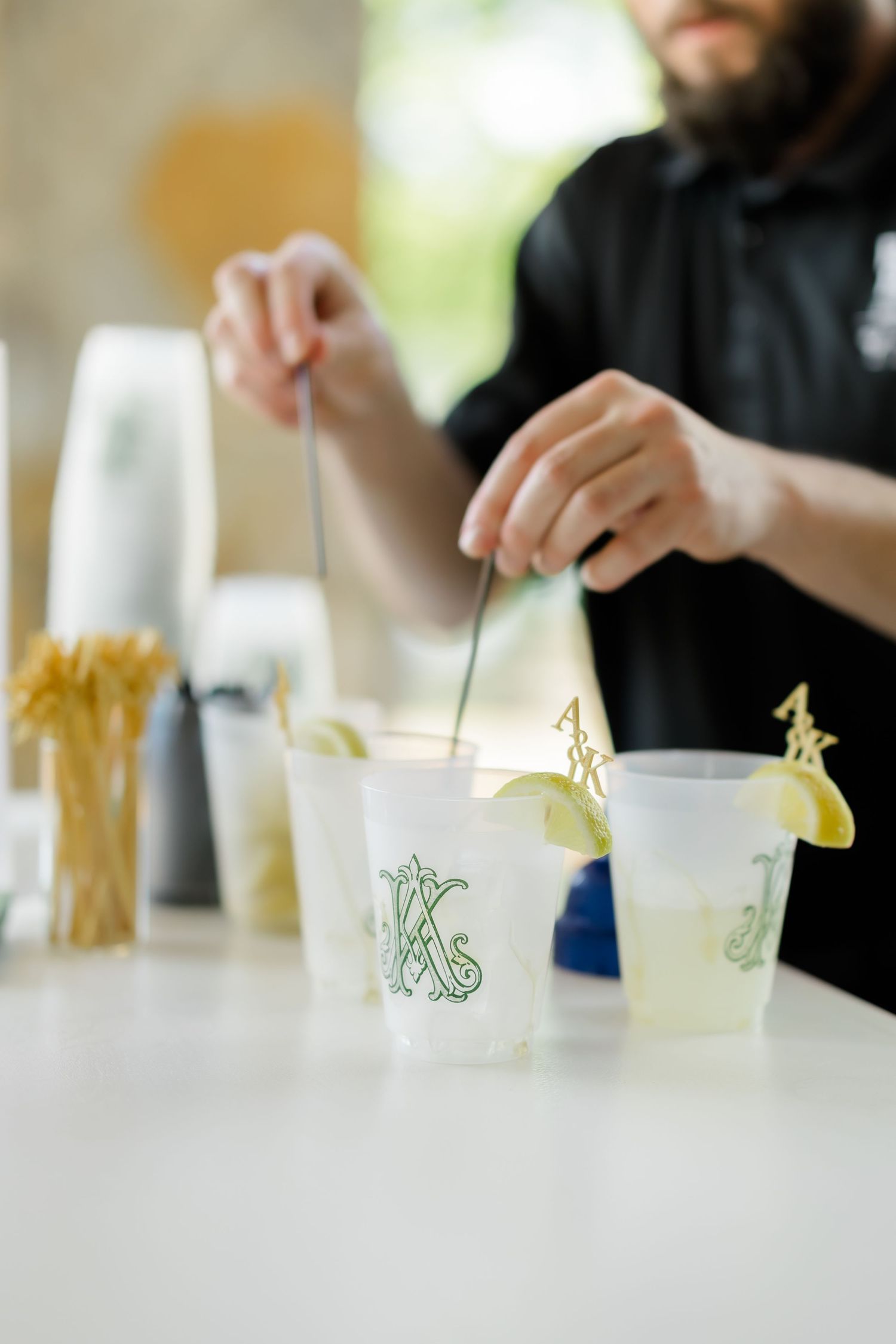 Bartender preparing cocktails in monogrammed frosted cups with lime garnishes and stirrers on a white bar counter.