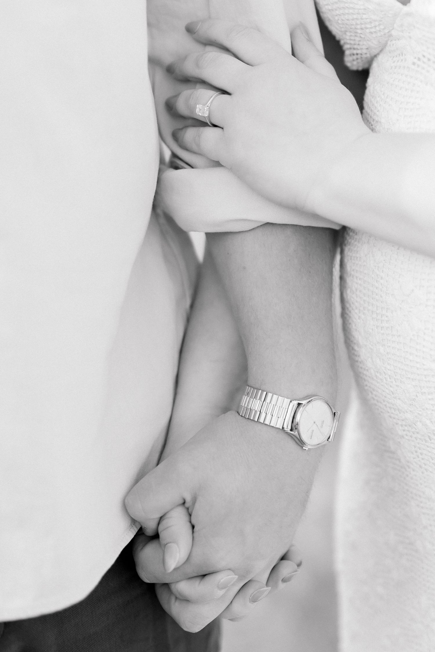 Close-up black and white photo of couple holding hands, woman wearing engagement ring and silver watch.