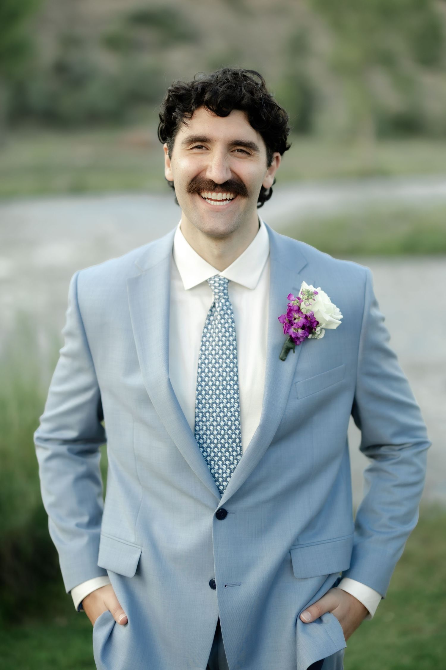 Smiling groom with mustache in light blue suit and floral boutonniere poses with hands in pockets outdoors.