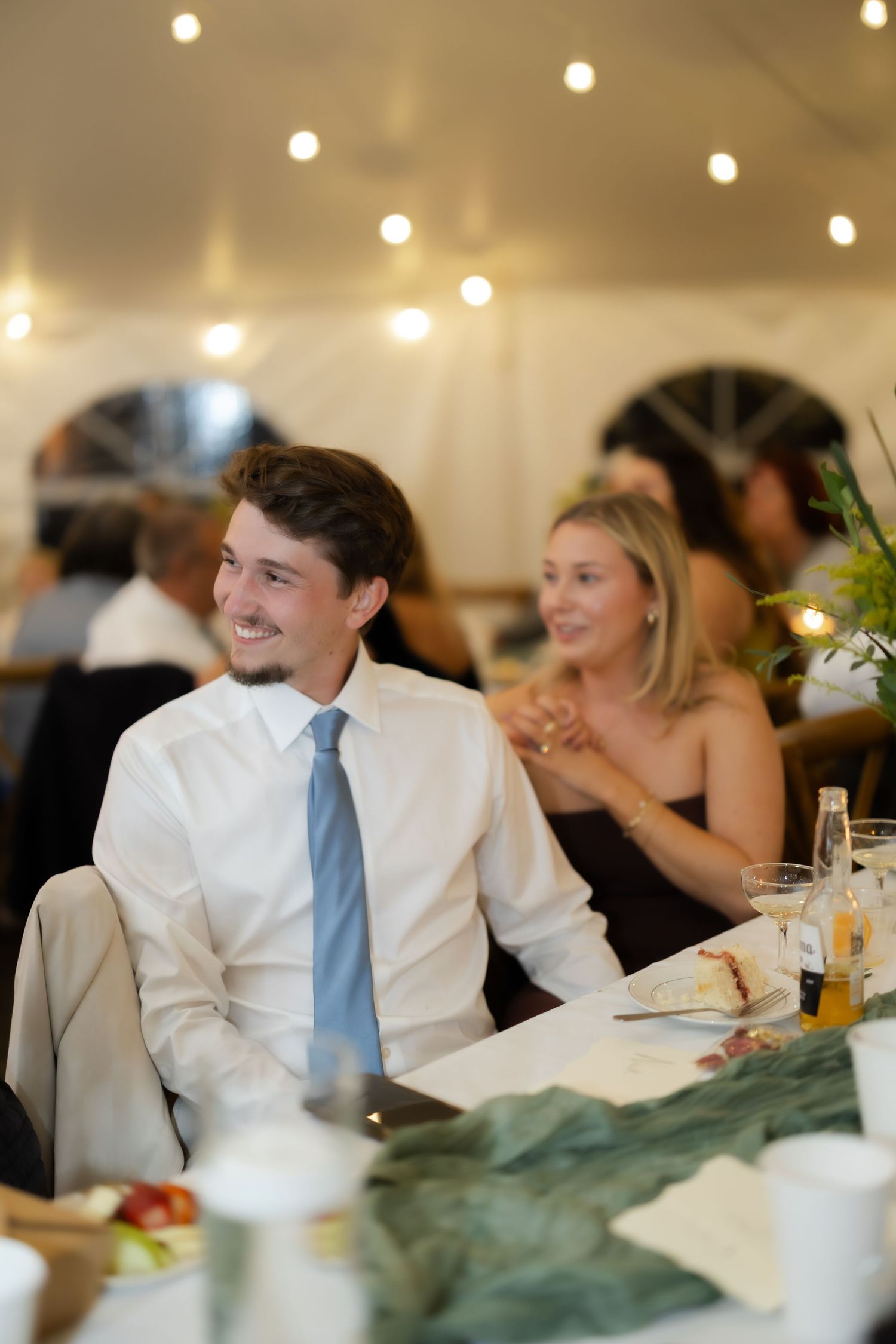 Young man in white shirt and blue tie smiling at wedding reception dinner table with string lights.