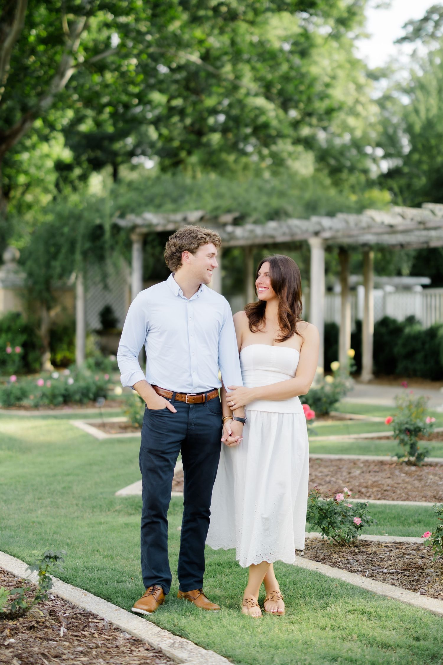 Couple holding hands and gazing at each other in a formal garden, man in light blue shirt, woman in white dress.