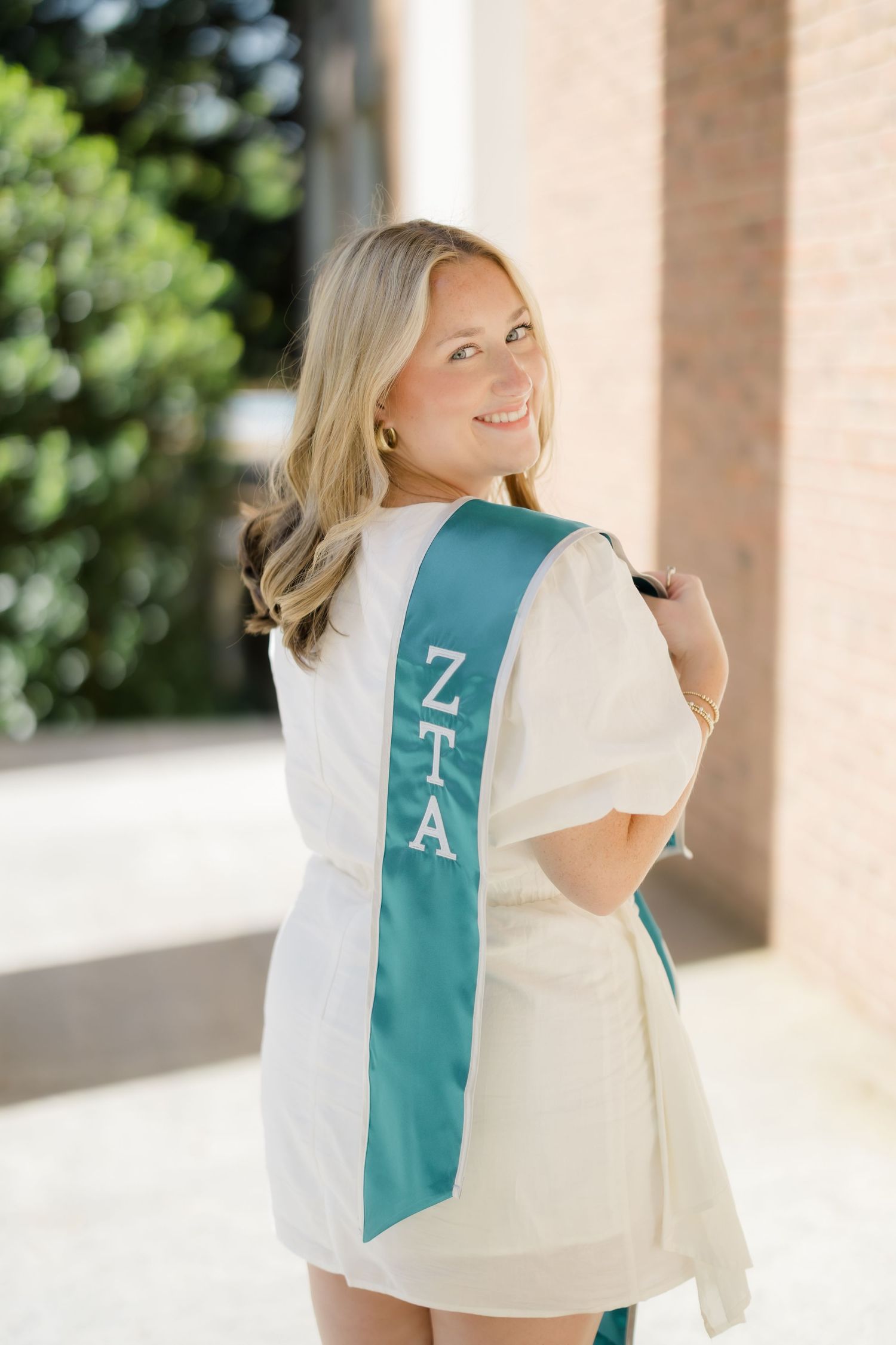 Smiling blonde woman wearing a white outfit and teal ZTA sorority sash, posing outdoors near a brick building.
