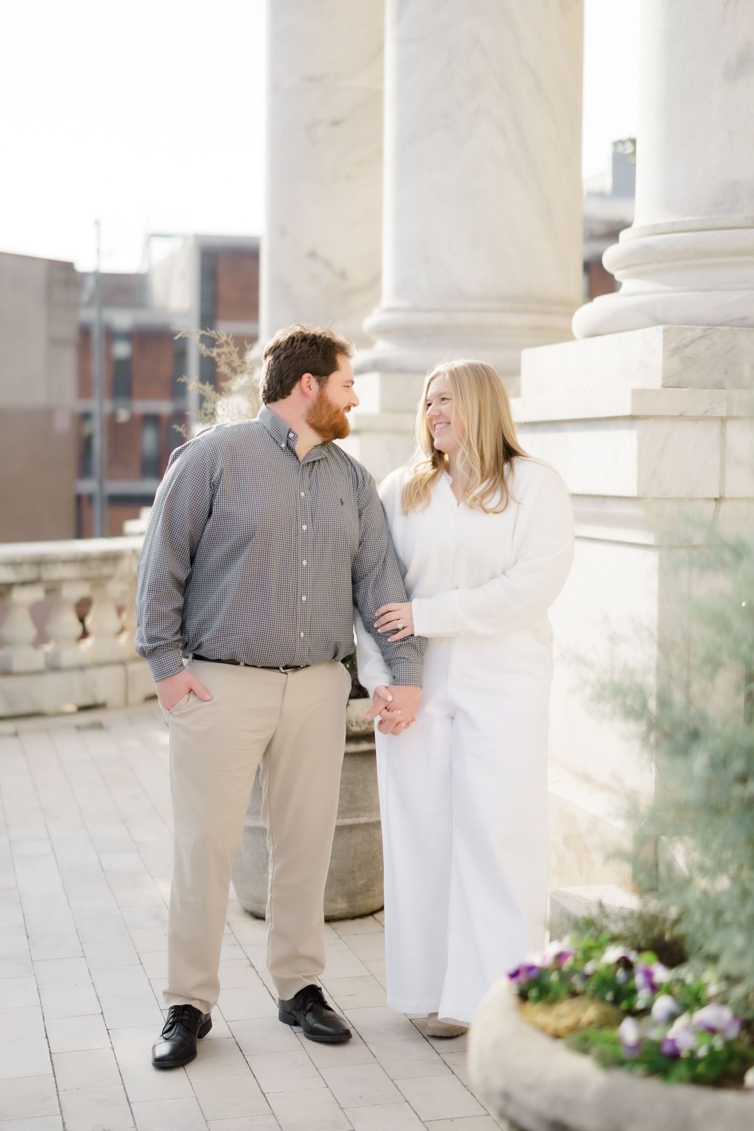 A couple smiles at each other on a marble terrace, man in gray shirt and khakis, woman in white outfit.