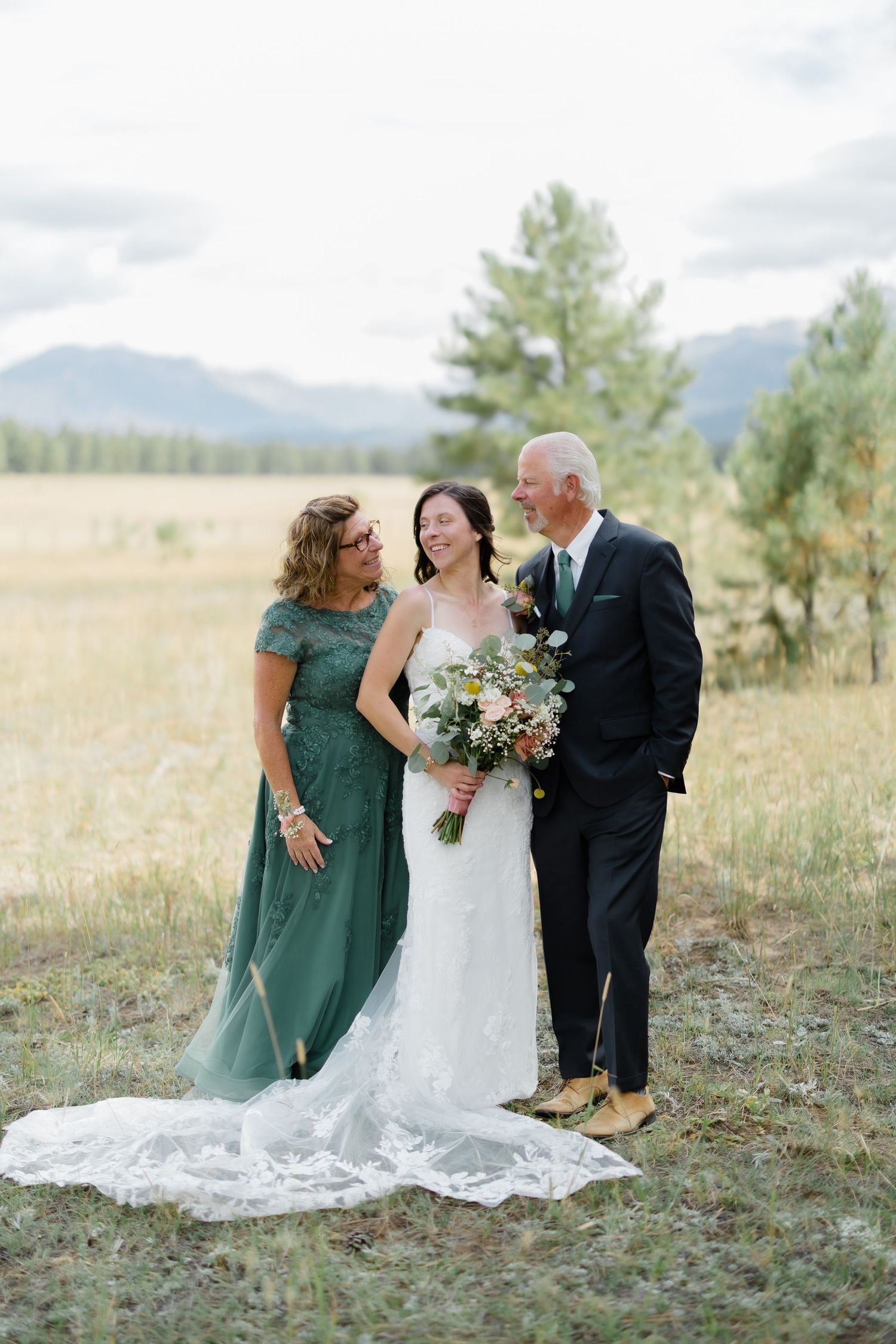 Bride in white lace gown holding bouquet stands with parents in green dress and dark suit at outdoor mountain wedding.
