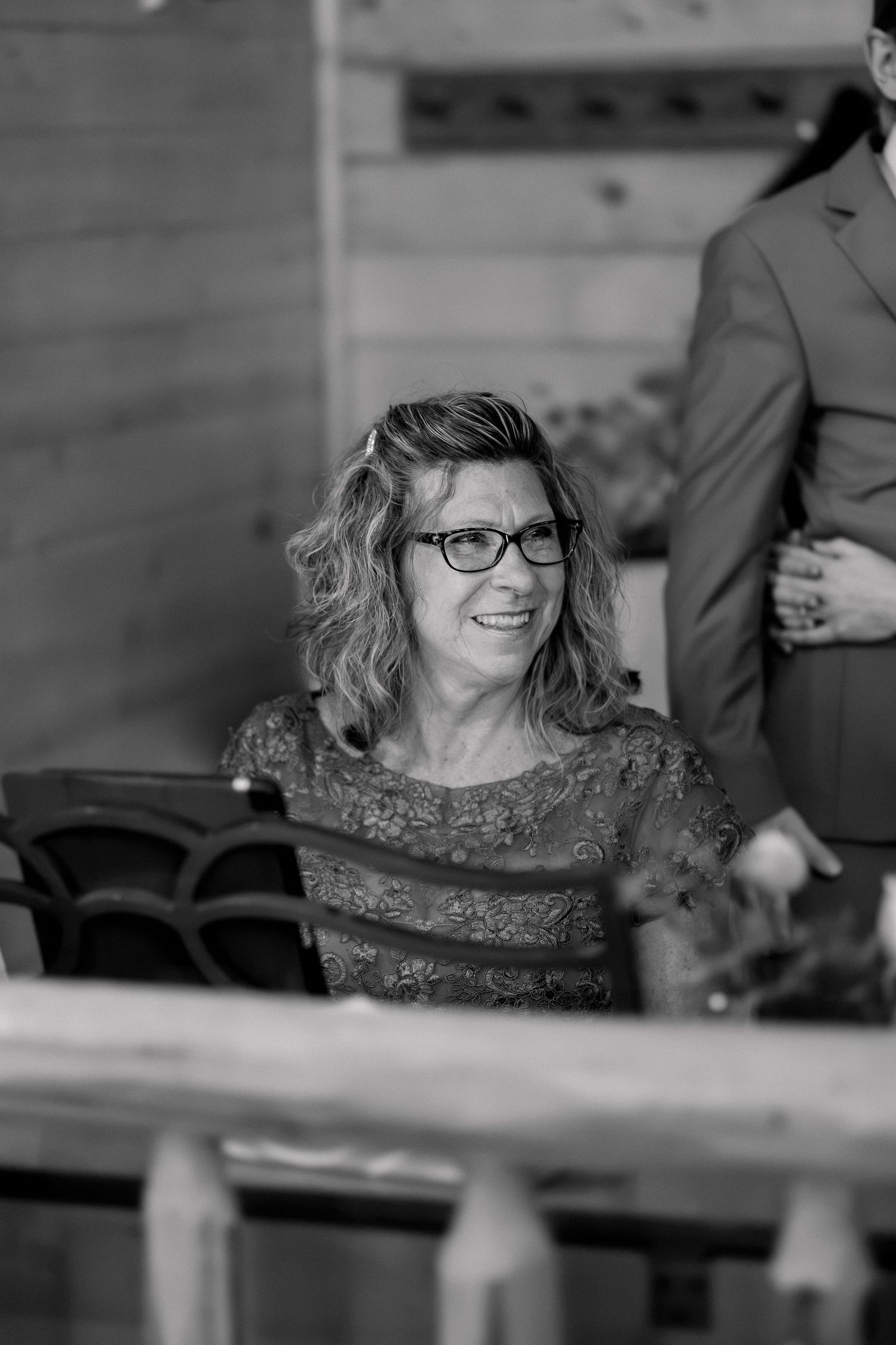 Black and white photo of a smiling woman with glasses and curly hair seated at a wedding ceremony.
