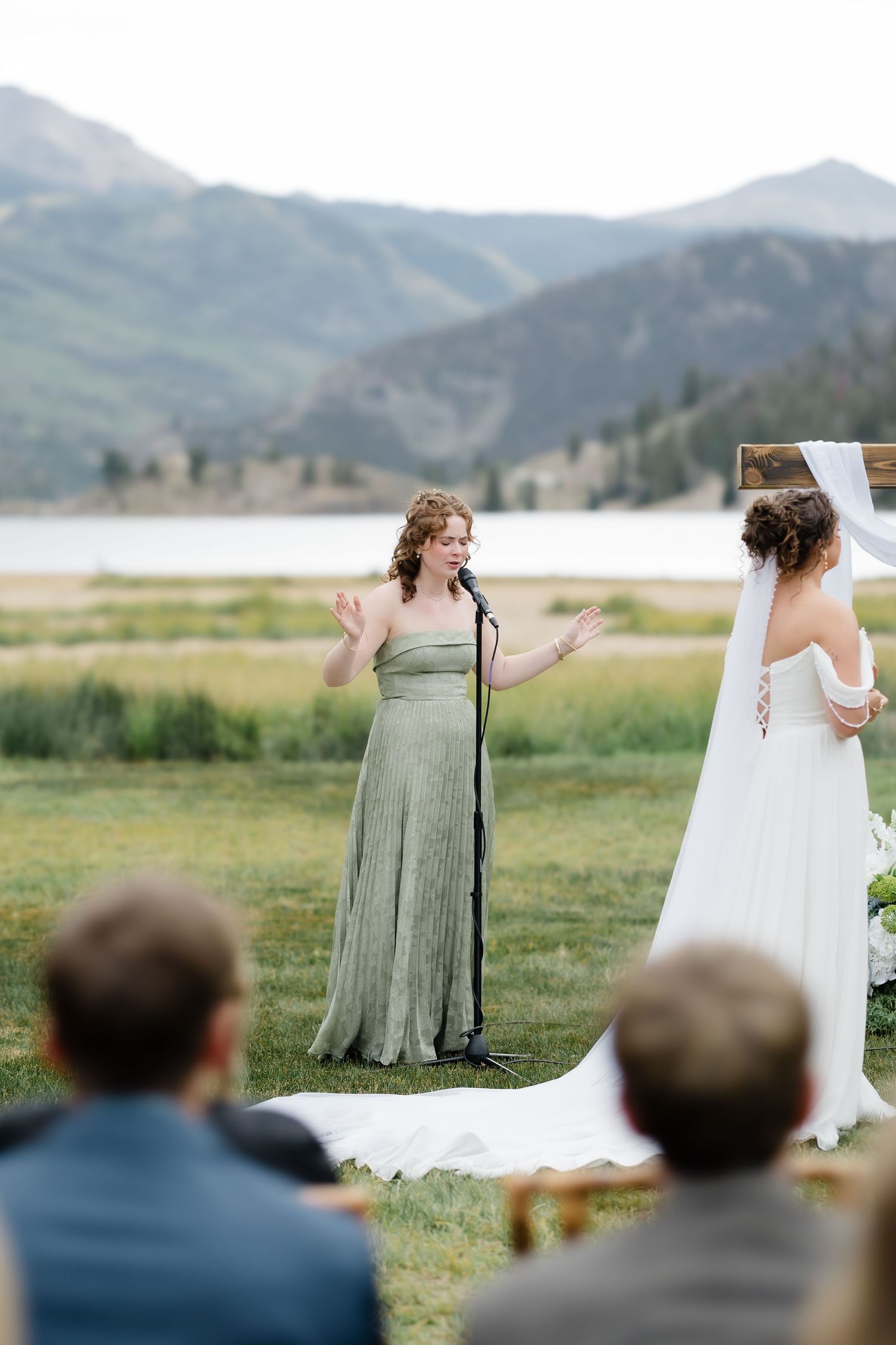 Woman in sage green gown speaking at microphone during outdoor lakeside wedding ceremony with mountains.