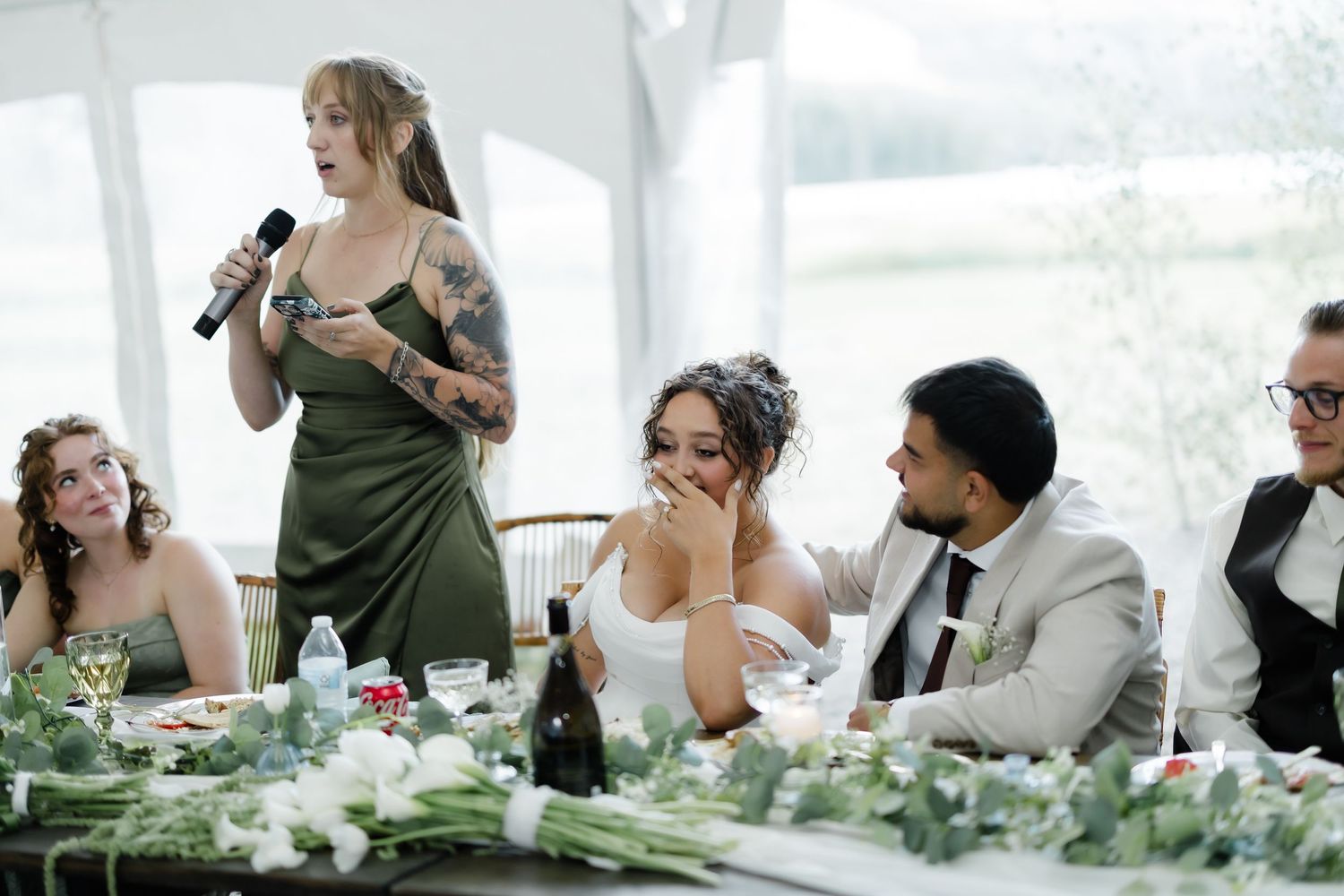 Bridesmaid in green dress gives wedding toast speech while bride and groom smile at reception table.