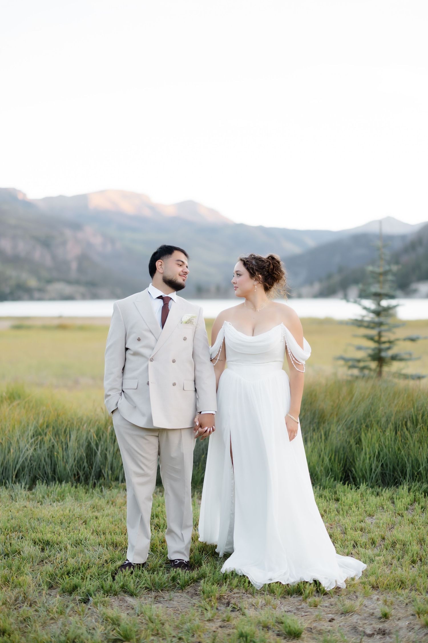 Bride and groom holding hands in a mountain meadow, she in a white off-shoulder gown, he in a beige suit.