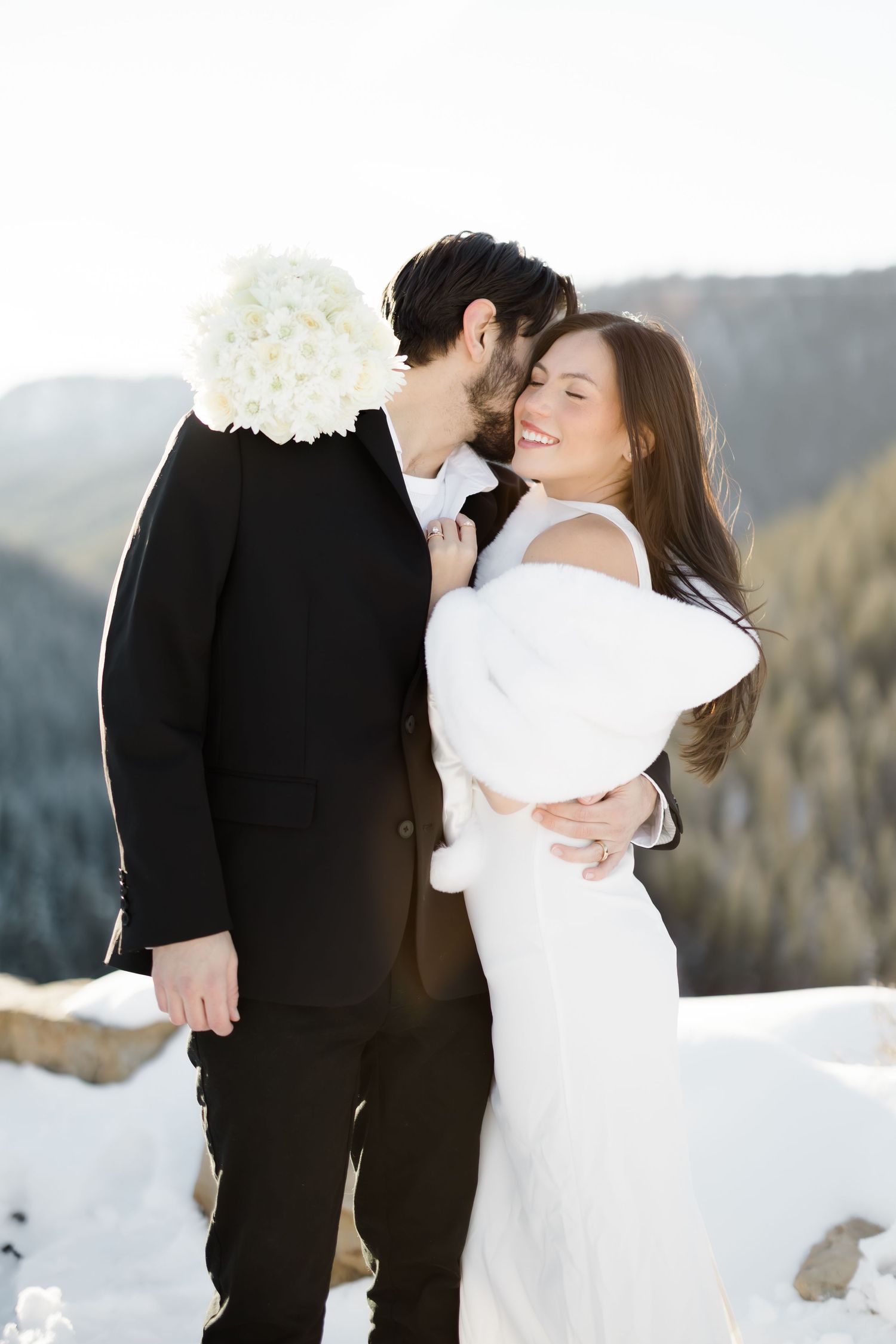 Happy bride smiles as groom kisses her cheek during winter mountain wedding photo session.