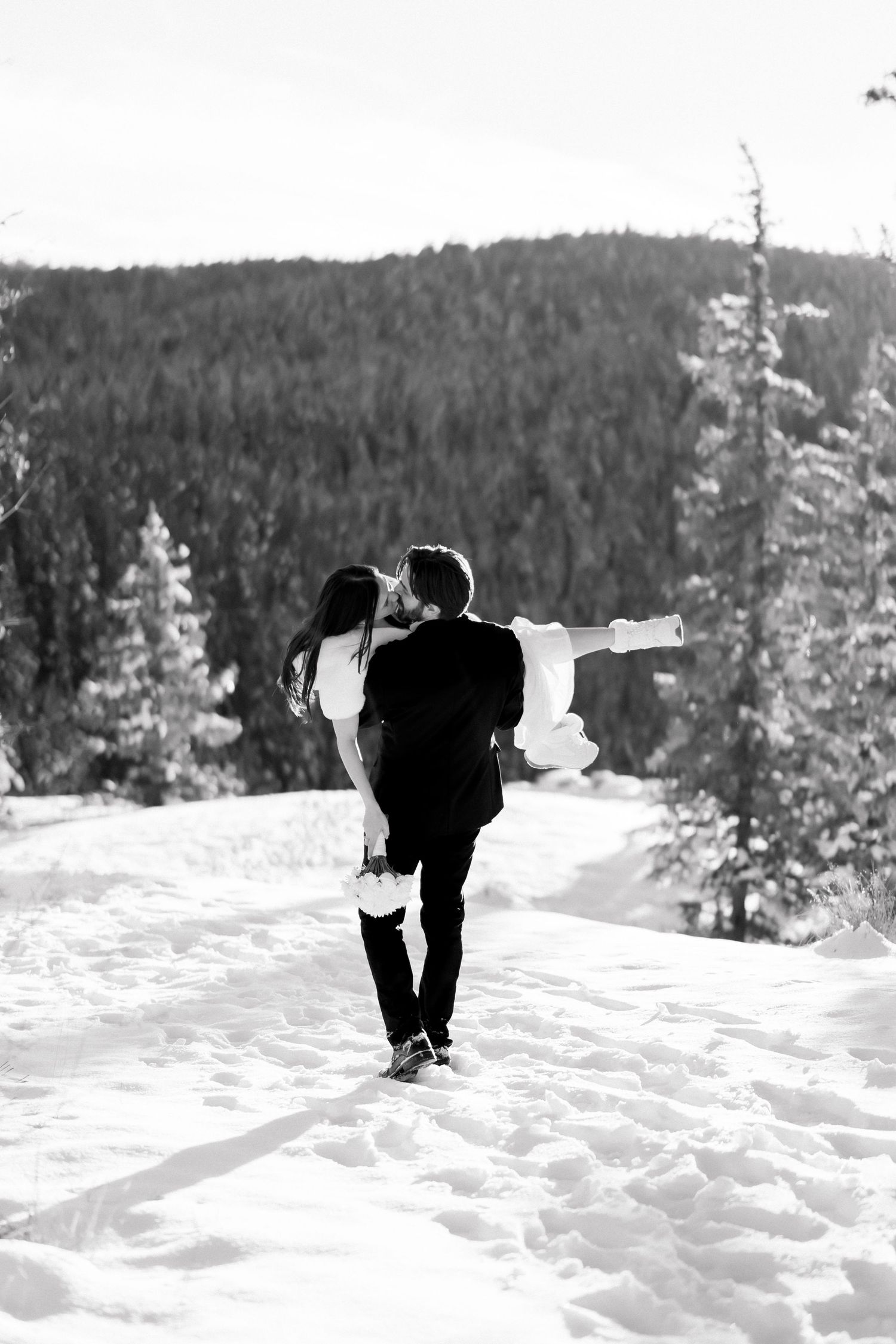 Black and white photo of groom carrying bride through snowy mountain landscape while they share a kiss.