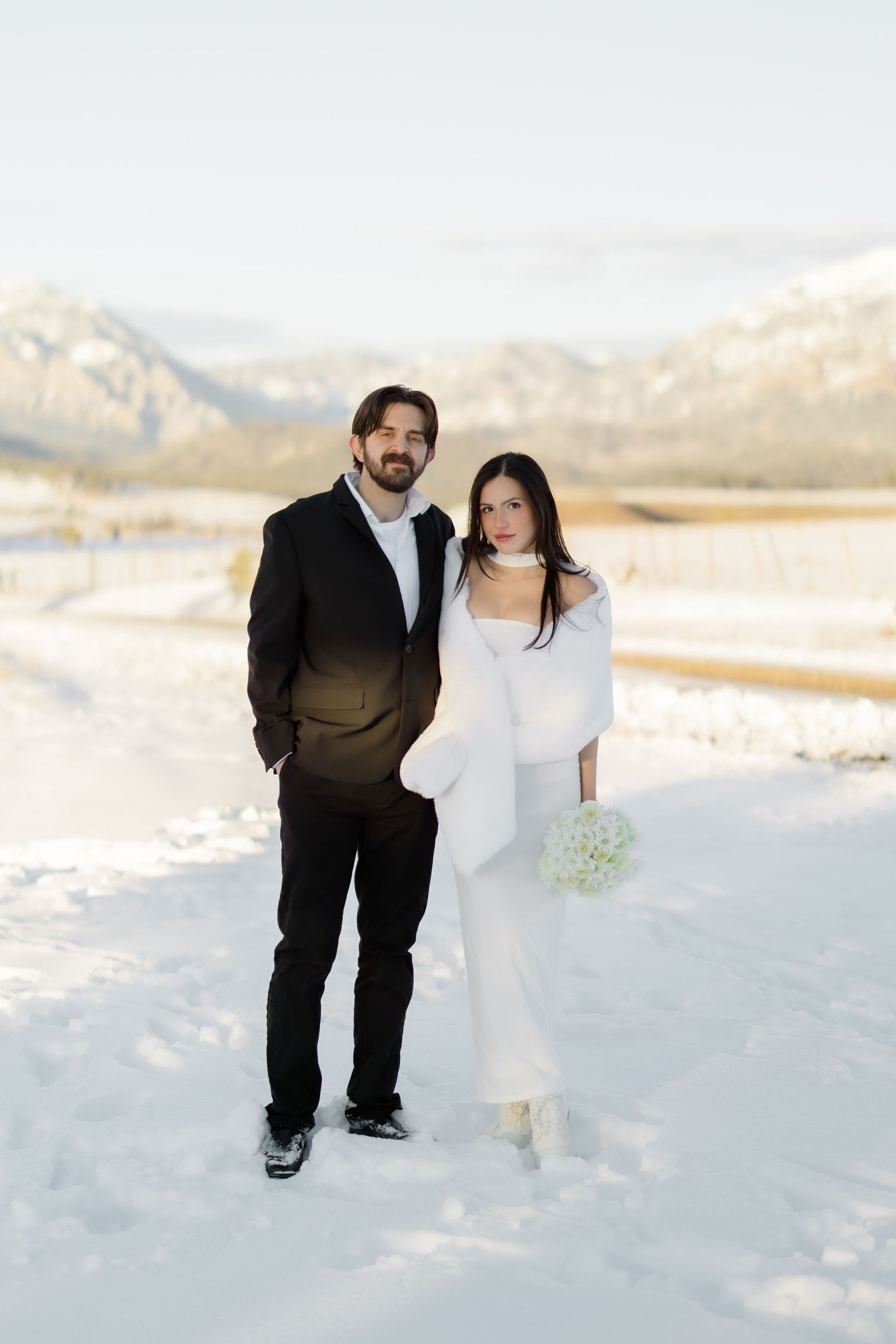 Bride and groom stand together in snowy mountain landscape, she holds white bouquet wearing fur wrap.