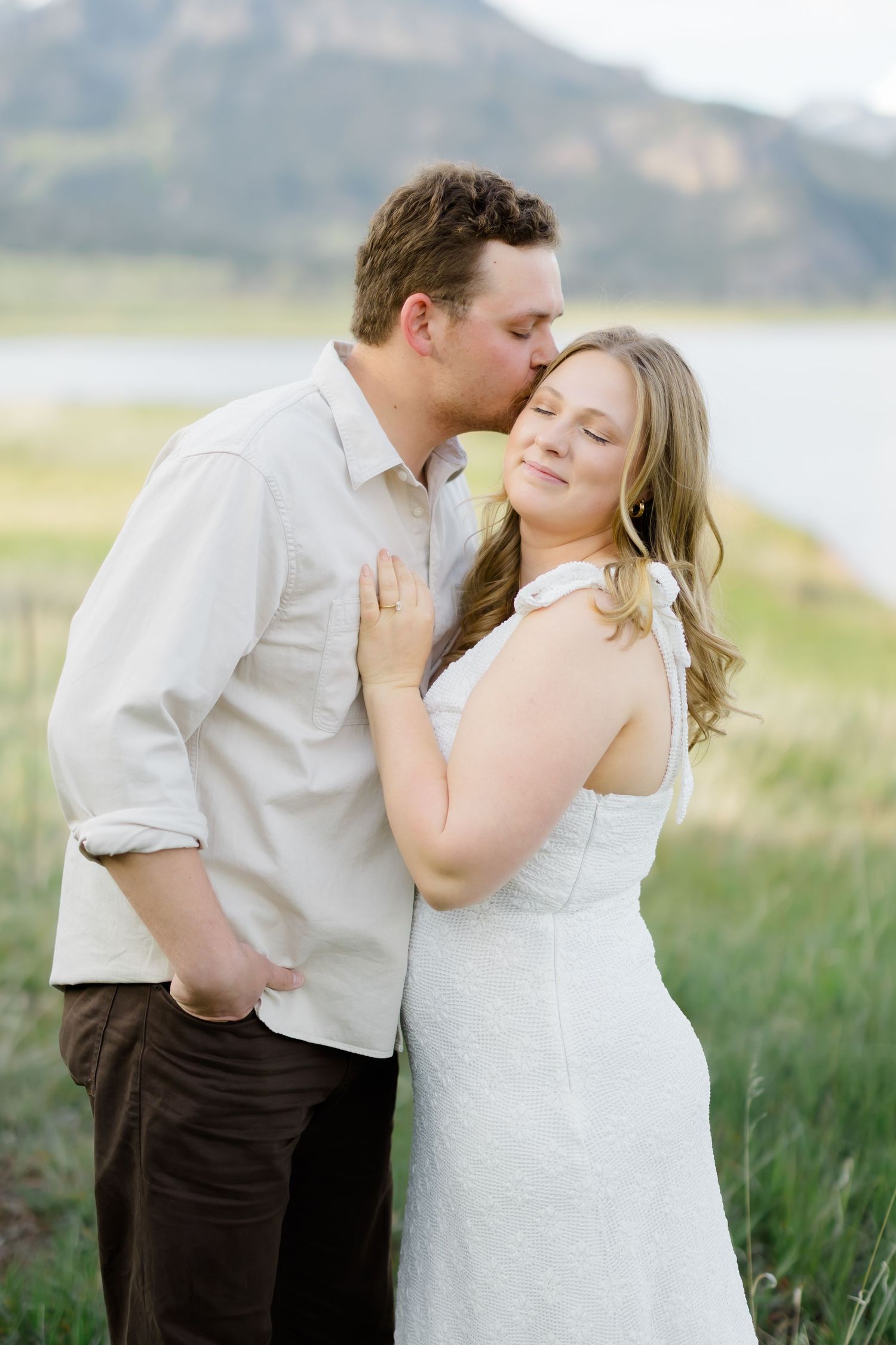Couple sharing a tender moment outdoors, man kissing woman's temple near a mountain lake.