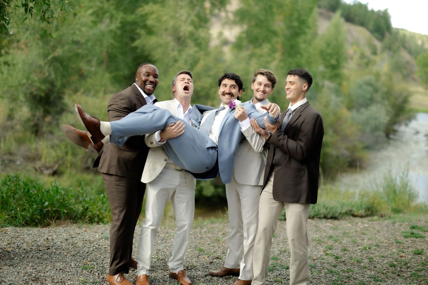 Groomsmen laughing while carrying groom in blue suit outdoors at wedding near green trees and river.