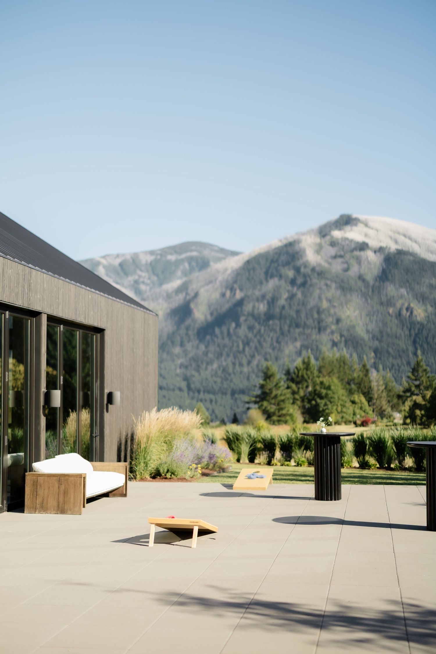 Modern patio with wood lounge chair and cornhole game, overlooking lush gardens and dramatic mountain views under clear blue sky.