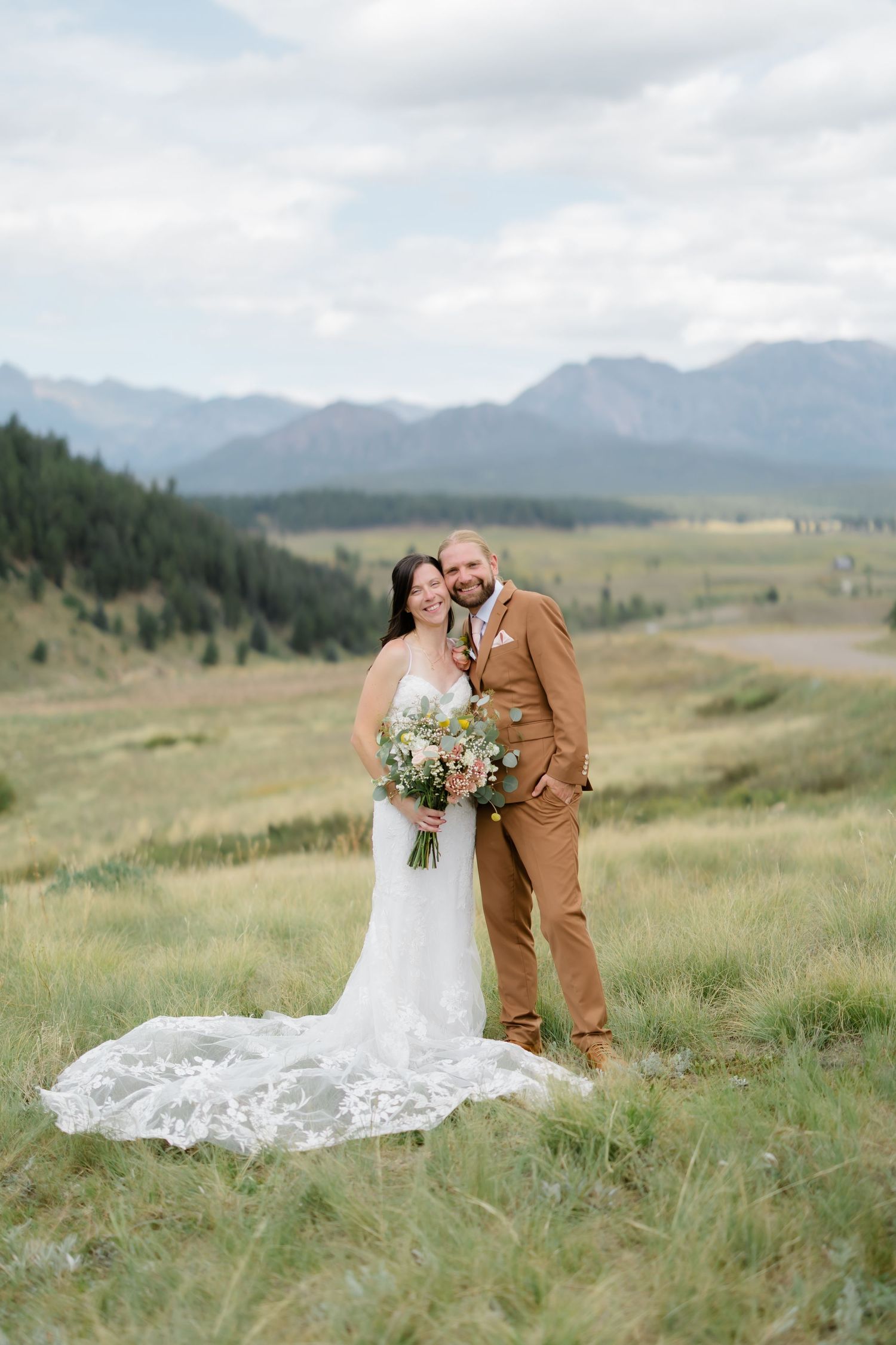 Bride and groom pose in mountain meadow, she in lace gown with train, he in tan suit, holding wildflower bouquet.