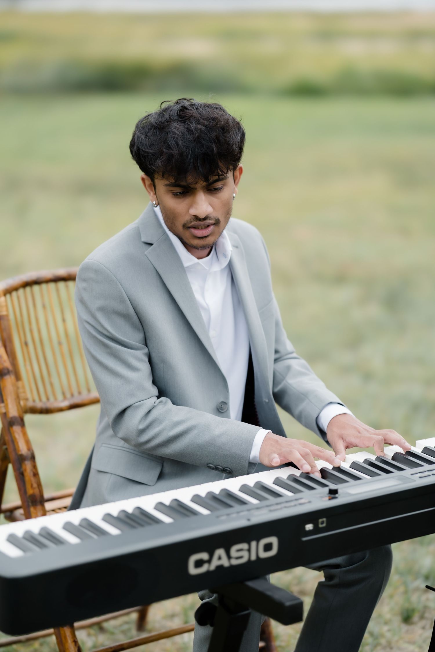 Young man in gray suit playing Casio keyboard piano outdoors in green field setting.