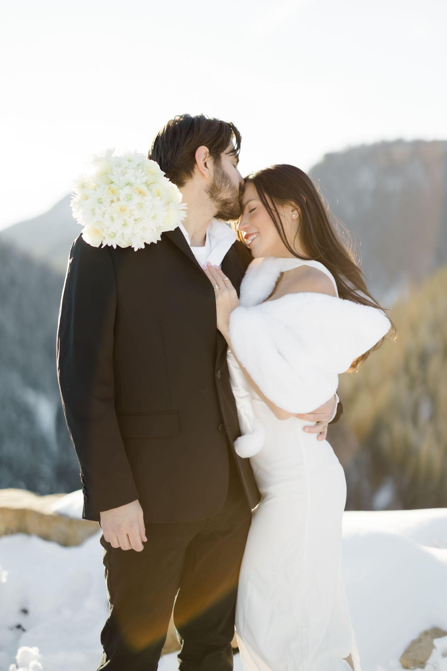 Bride and groom embrace in snowy mountain setting, she wears white fur wrap holding bouquet.