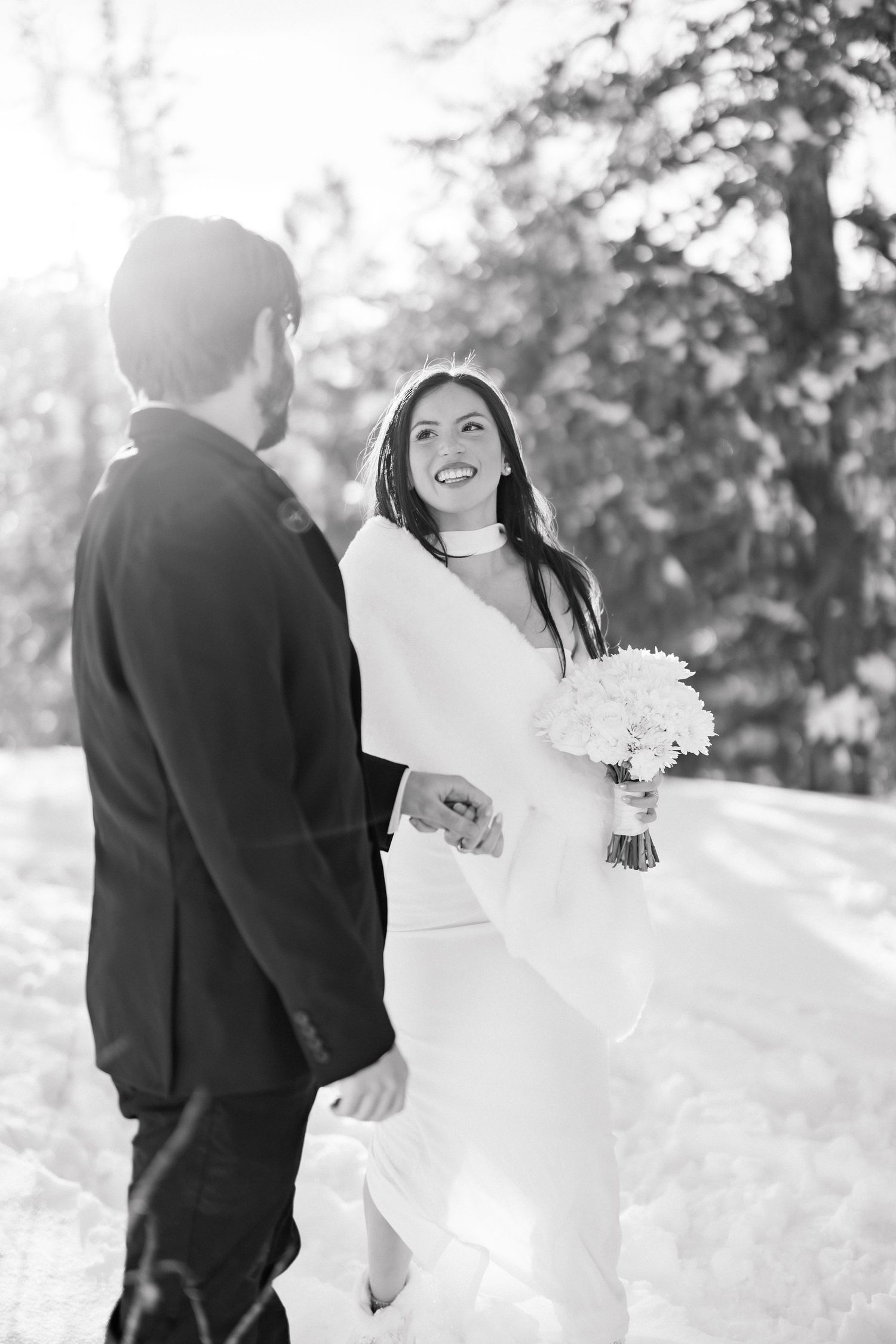 Black and white photo of laughing bride holding bouquet walking hand in hand with groom through snowy forest.
