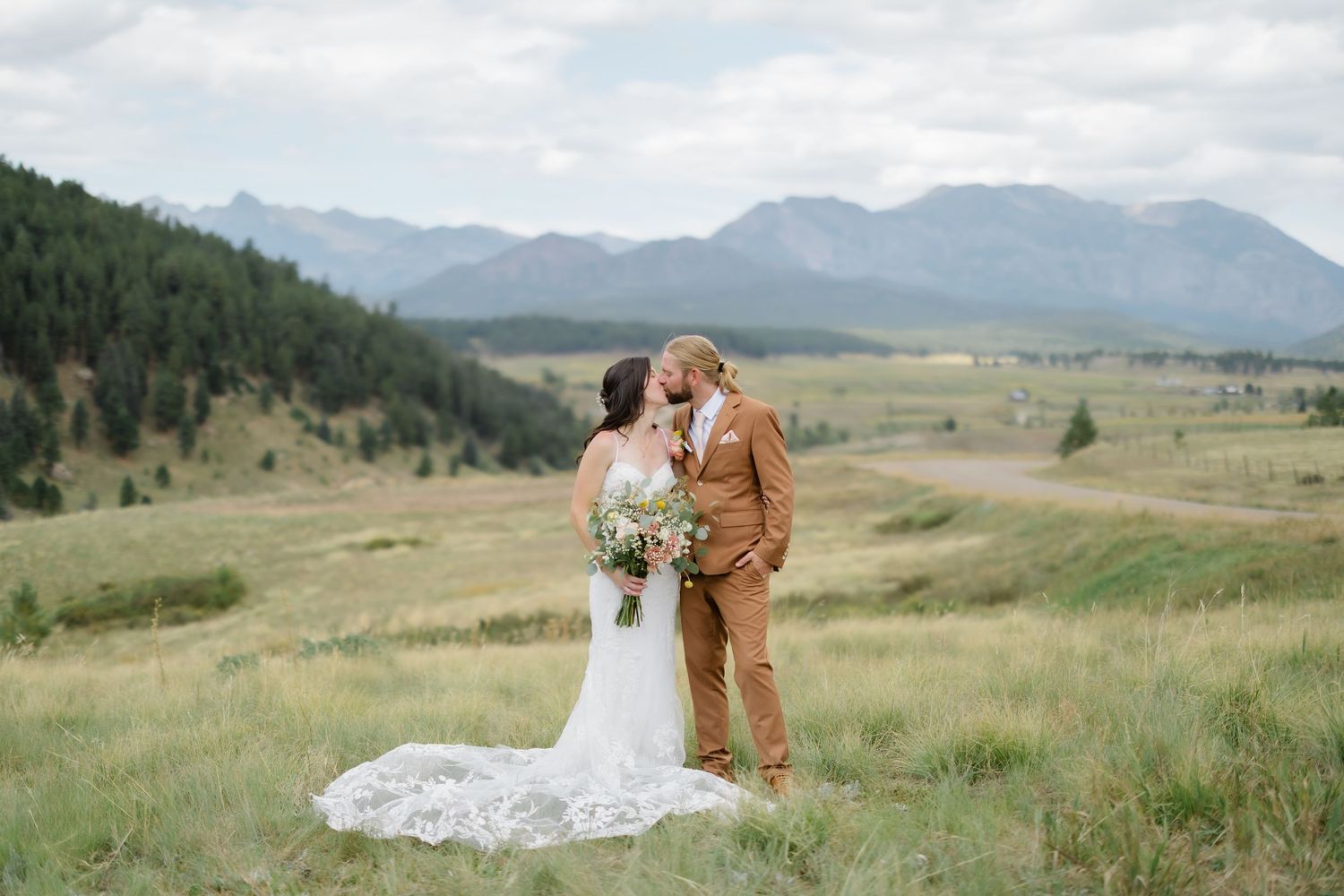 Bride and groom share a kiss in a mountain meadow, with stunning Rocky Mountain peaks and green forests in the background.