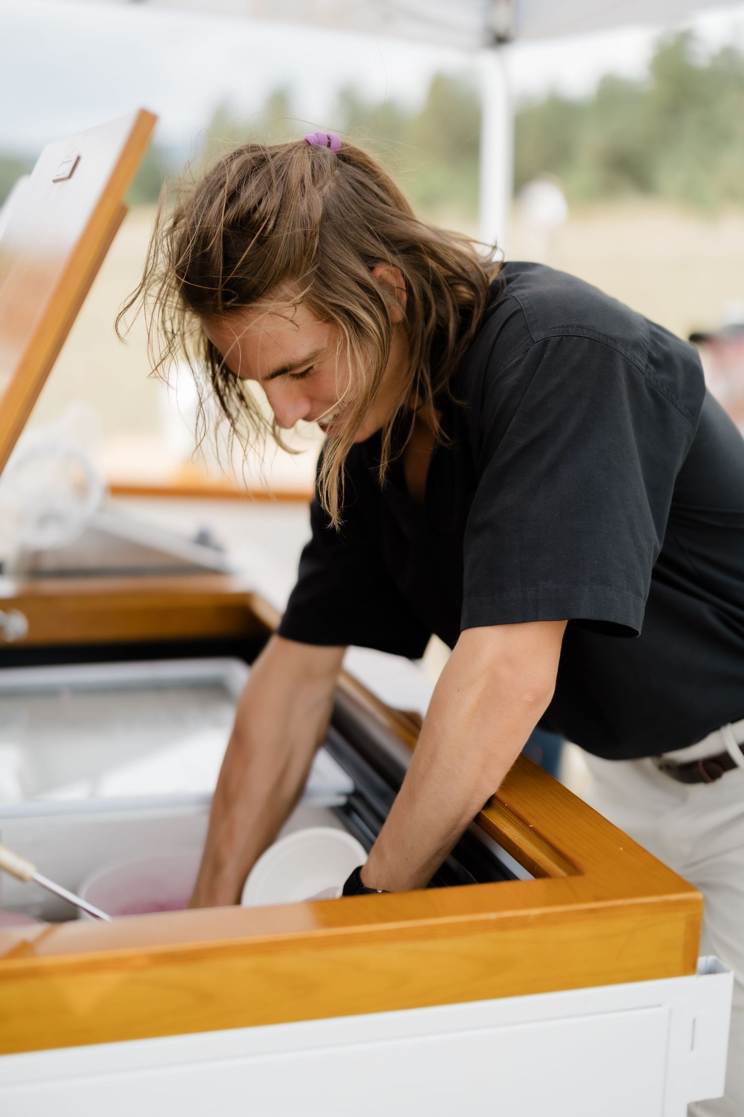 Long-haired man in black t-shirt leaning over a wooden piano outdoors at an event.