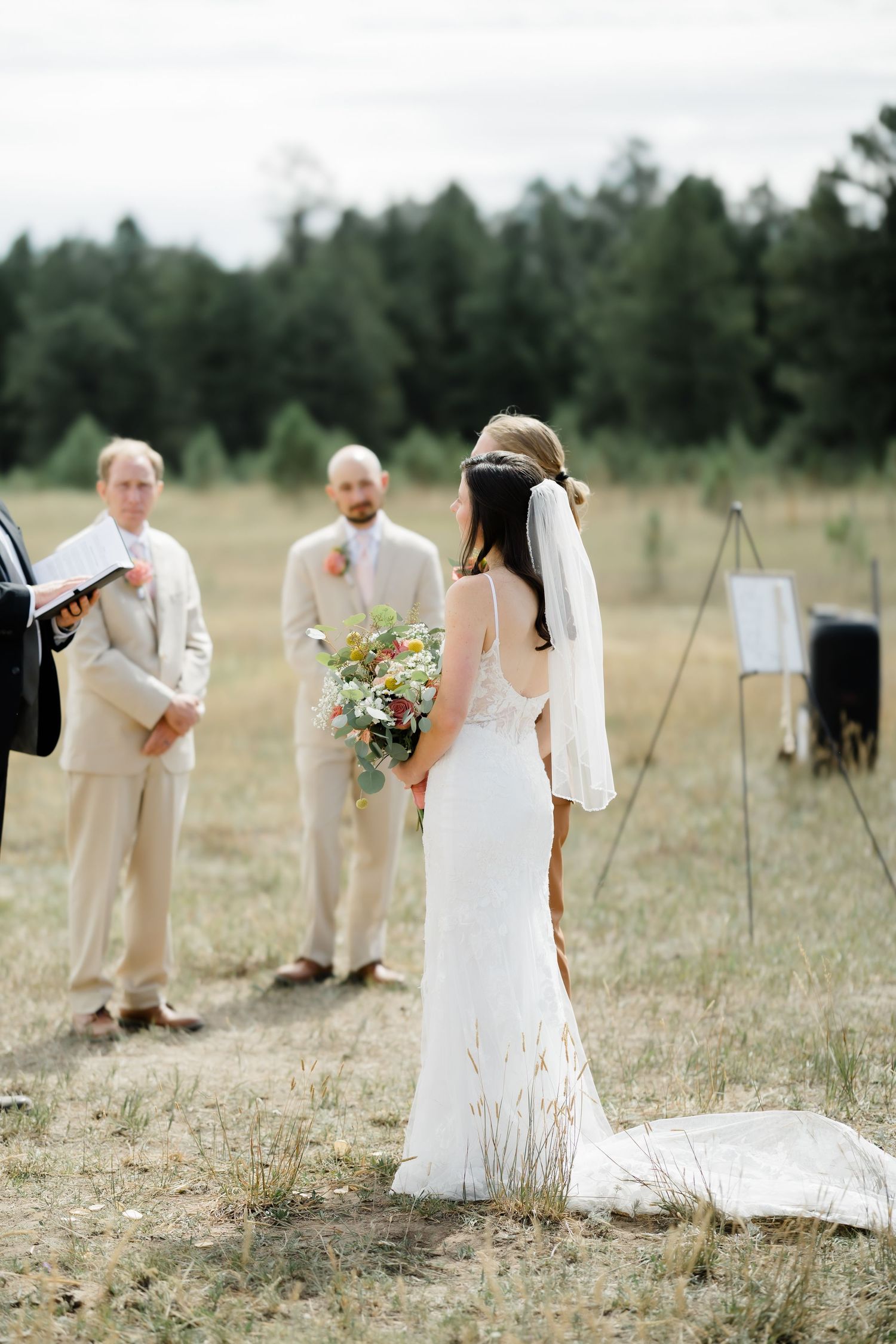 Bride in white gown holding bouquet stands before groom and officiant during outdoor wedding ceremony in grassy field.