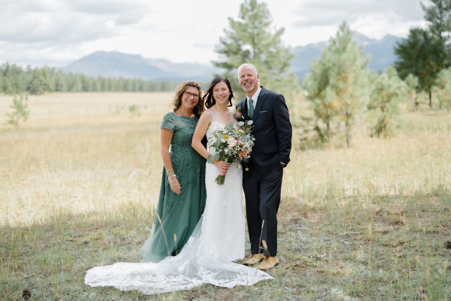 Bride in white gown with bouquet poses with family in green dress and suit against mountain meadow backdrop.