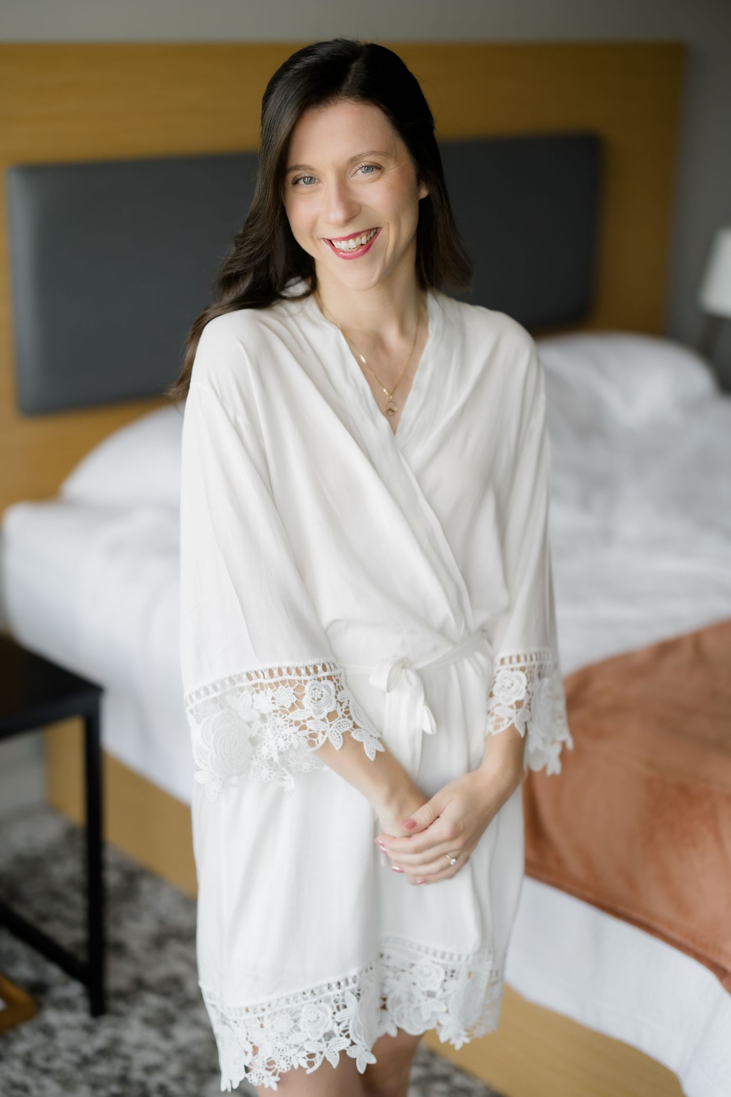 Smiling bride in white lace-trimmed robe standing in a hotel bedroom before her wedding.
