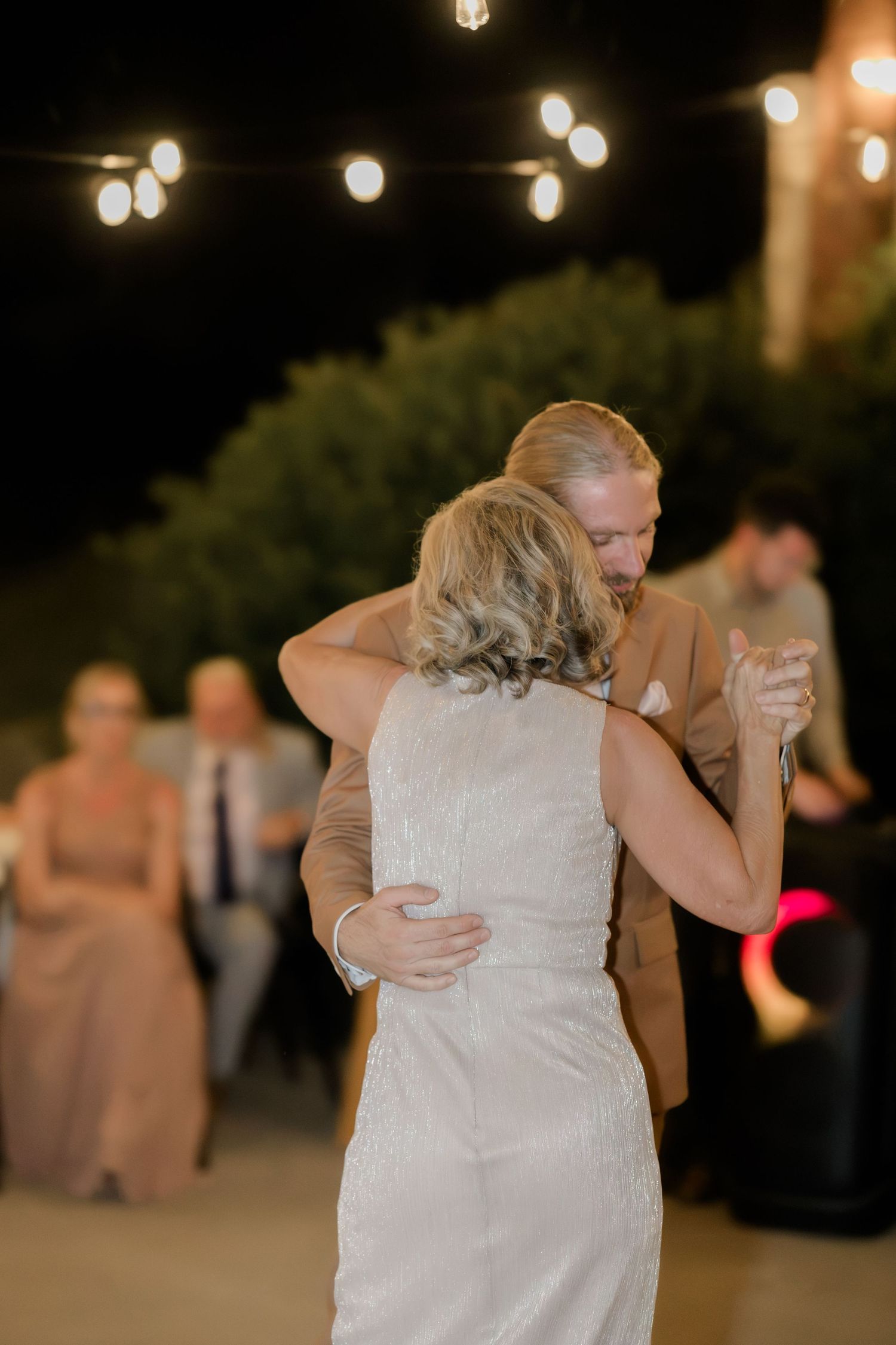 Bride and groom share an emotional first dance outdoors at night under warm string lights at their wedding reception.