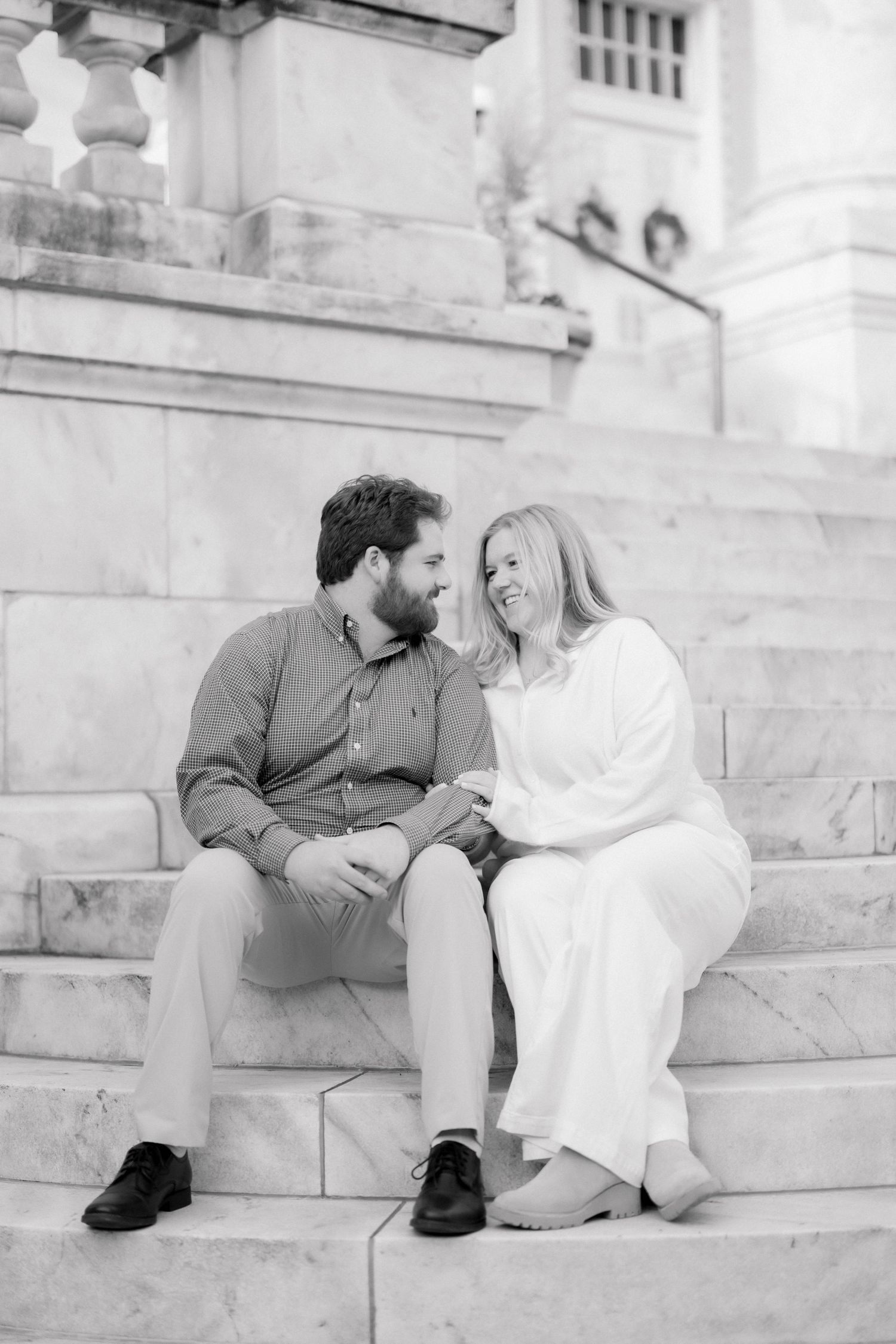 Black and white photo of a couple sitting on marble steps, smiling at each other during an engagement session.