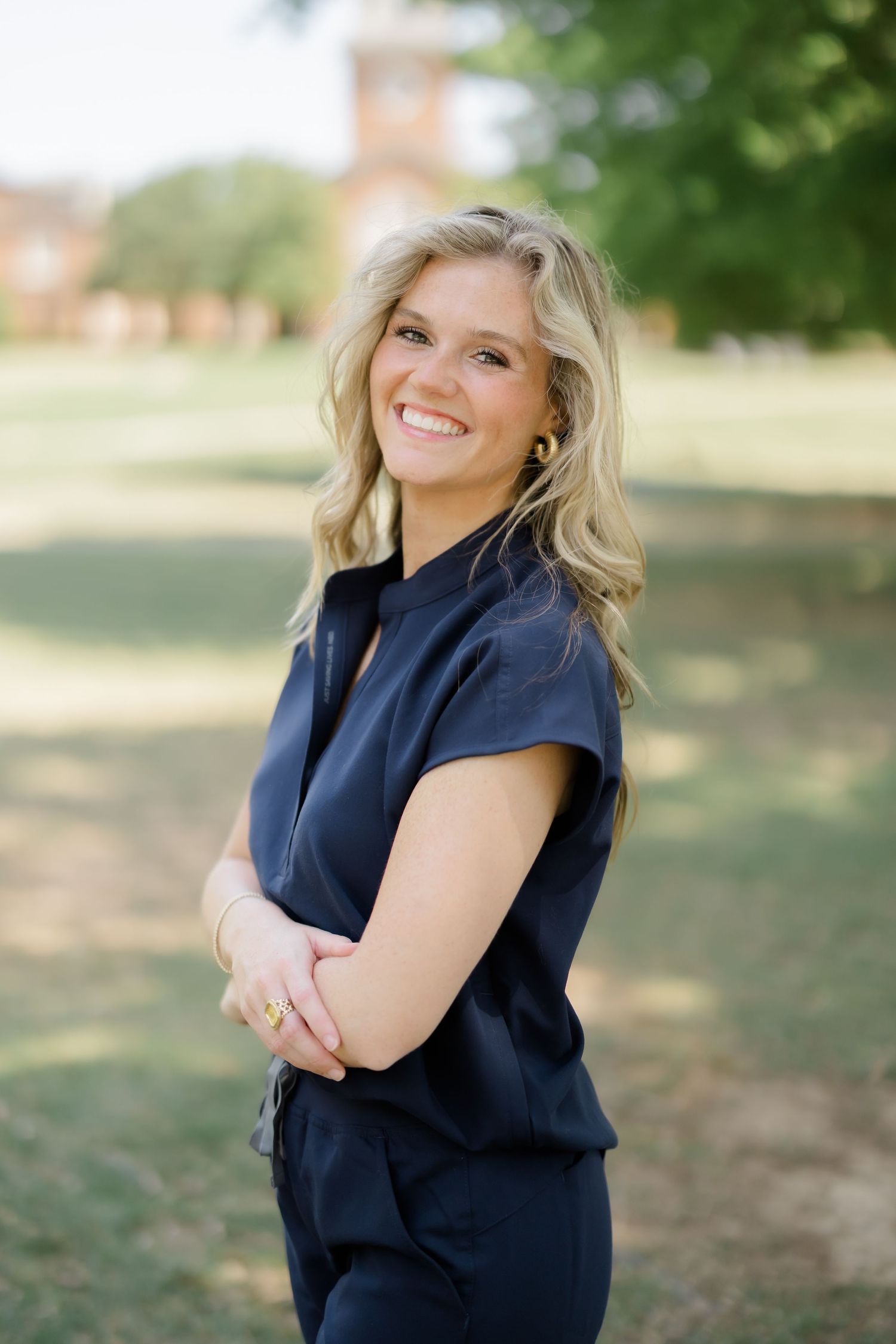 Smiling blonde woman in navy blue outfit standing outdoors on green campus grounds with brick building background.