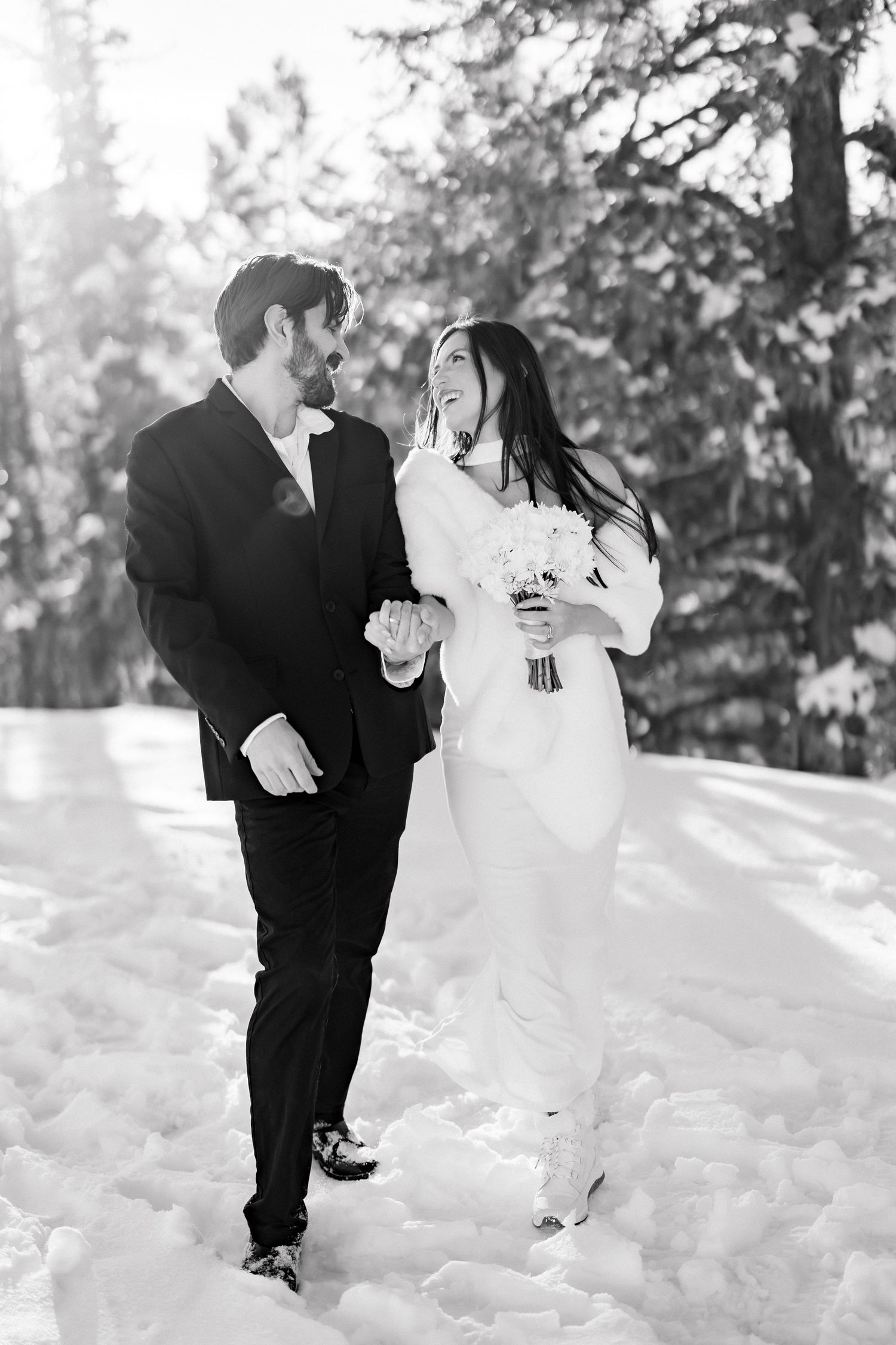 Bride and groom walk hand in hand laughing through snowy forest in black and white wedding photo.