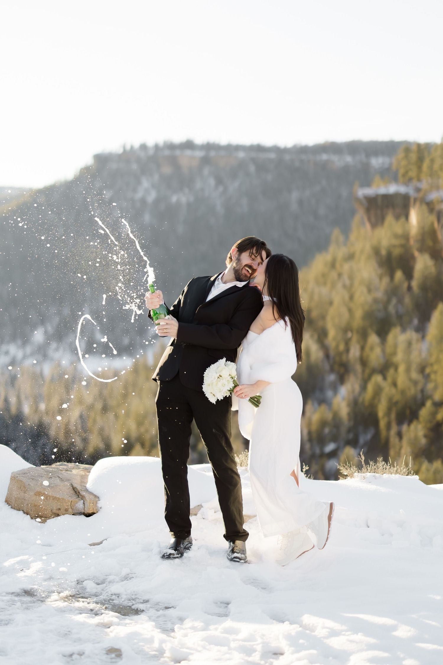 Newlyweds kissing while spraying champagne on a snowy mountain overlook in winter.