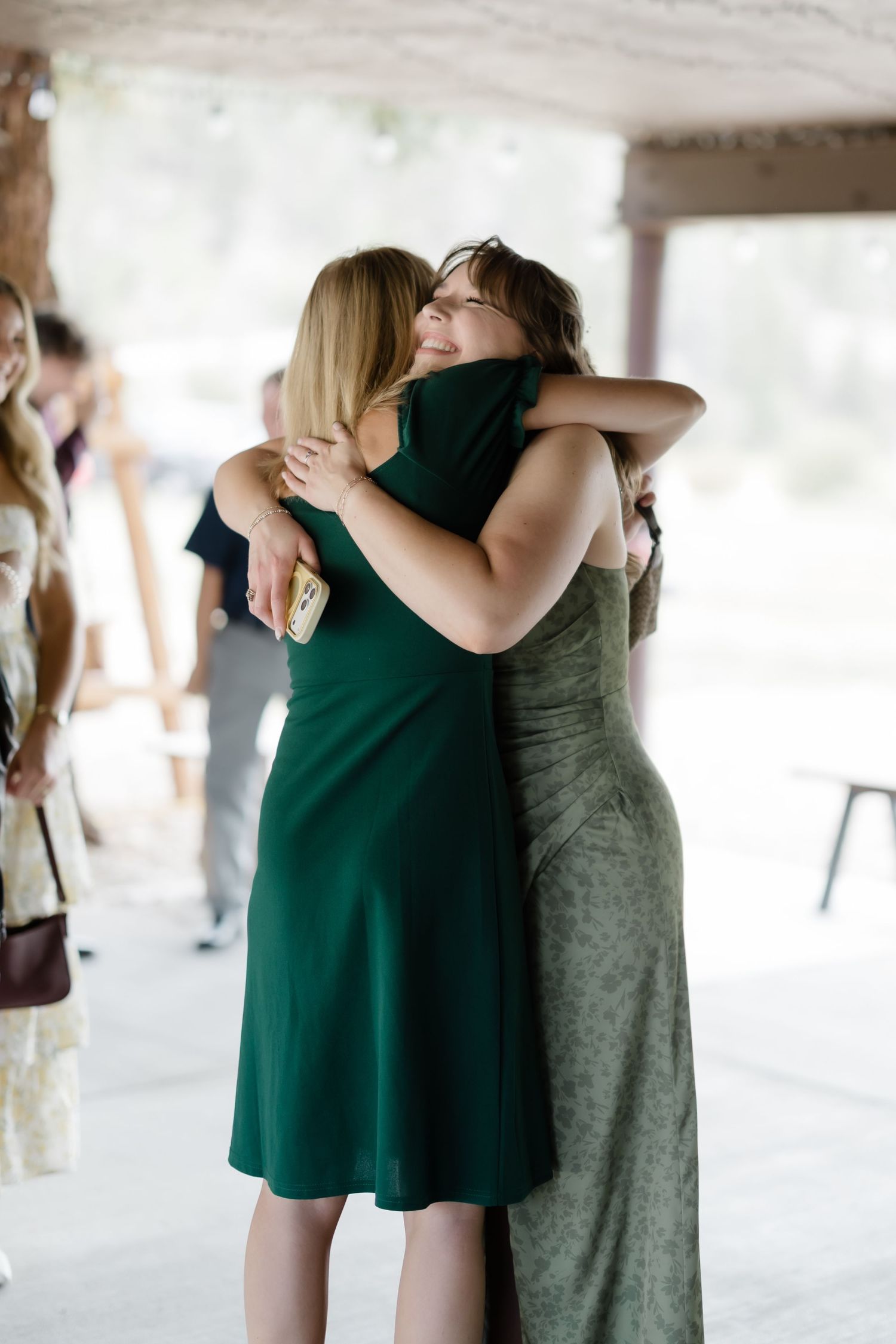 Two women in green dresses share a warm hug at an outdoor event or wedding celebration.