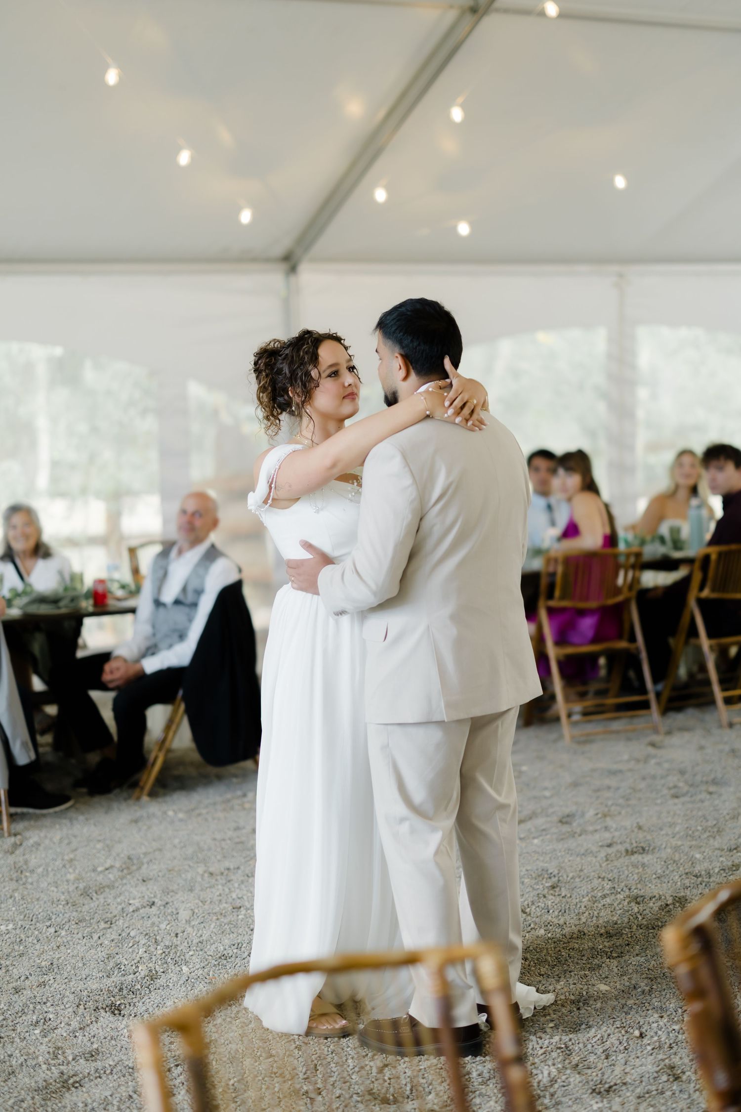 Bride and groom share first dance at tent wedding reception with seated guests watching.