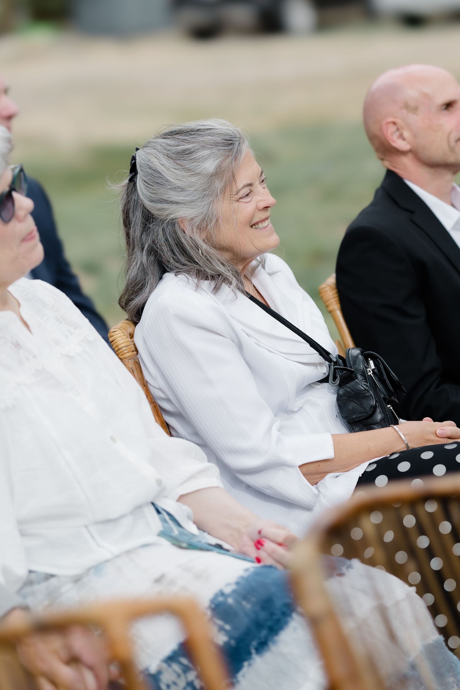 Smiling older woman with gray hair in white blouse seated outdoors at a wedding ceremony.