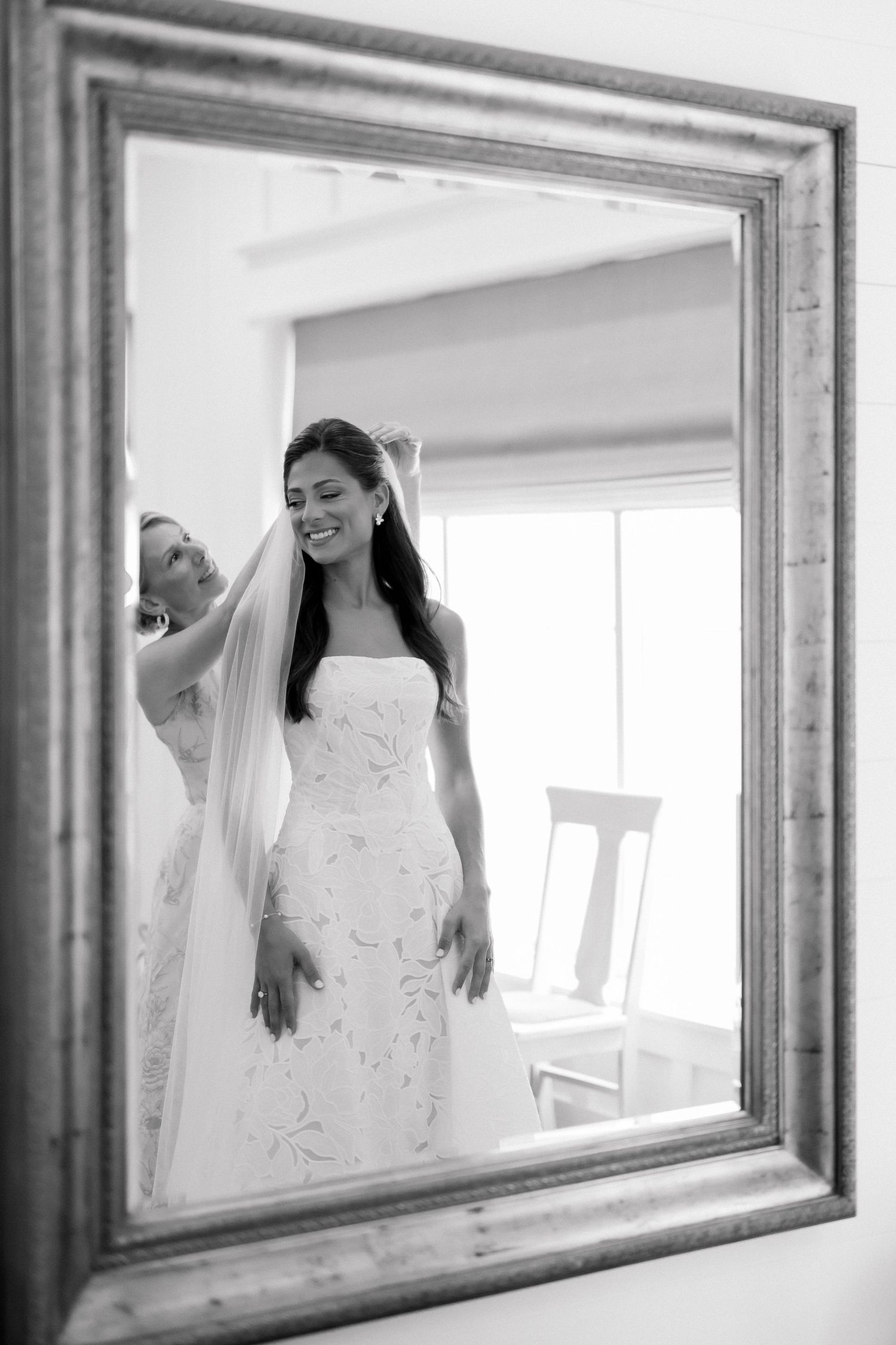 Black and white mirror reflection of a smiling bride in a floral gown having her veil adjusted by a bridesmaid.