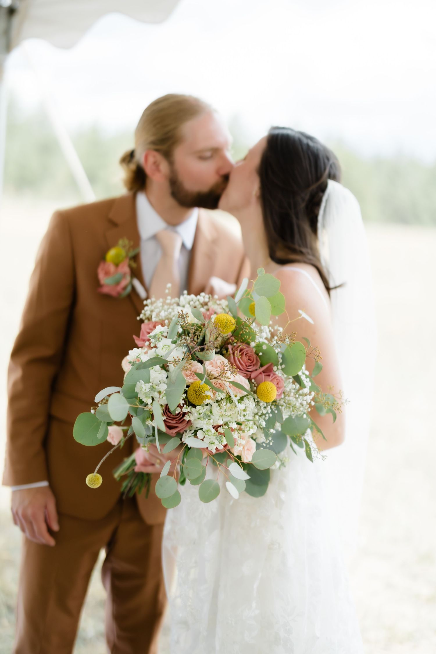 Bride and groom sharing a kiss at outdoor wedding, bride holding lush bouquet of roses, eucalyptus, and wildflowers.