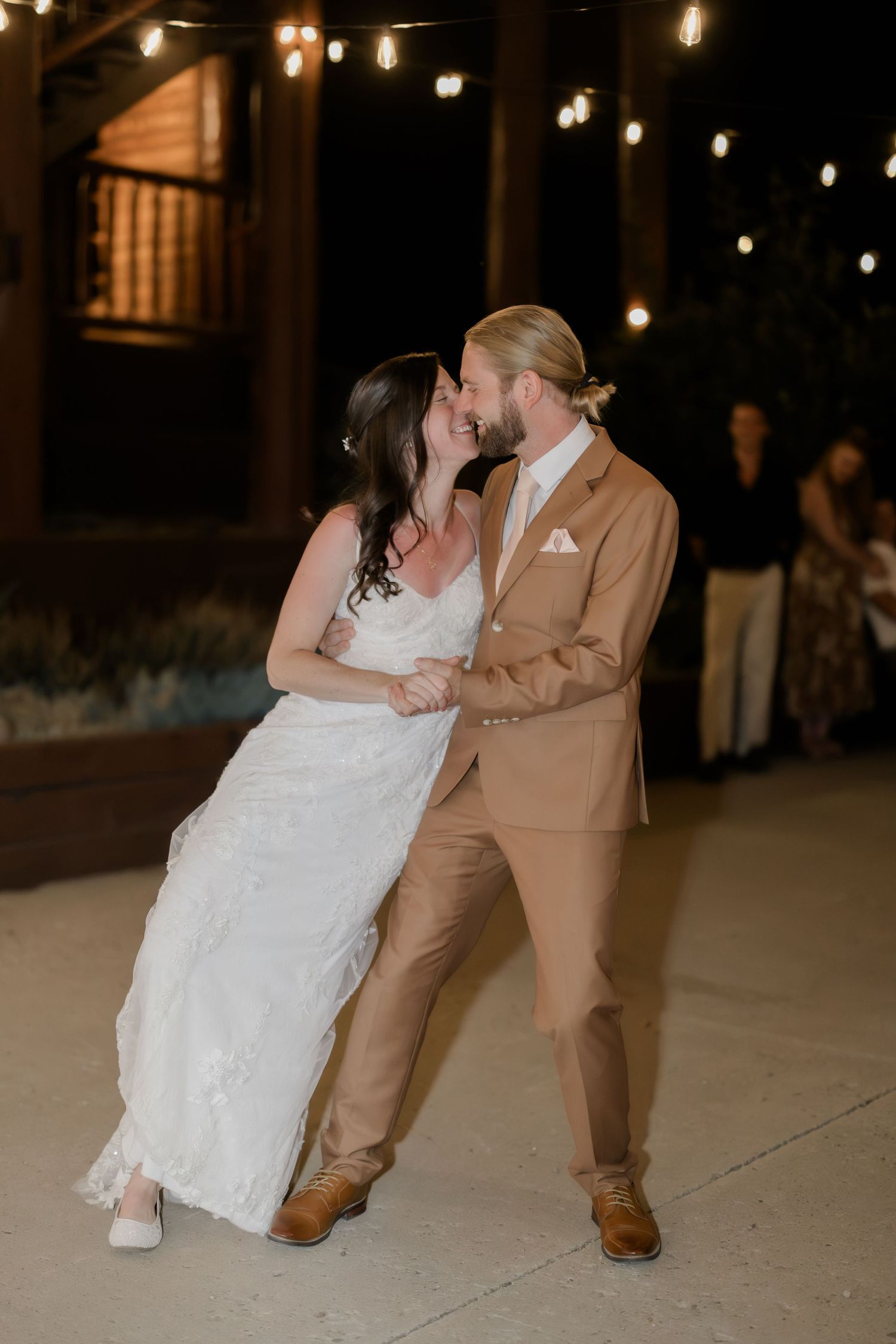 Bride and groom share first dance outdoors at night under string lights, bride in white gown, groom in tan suit.