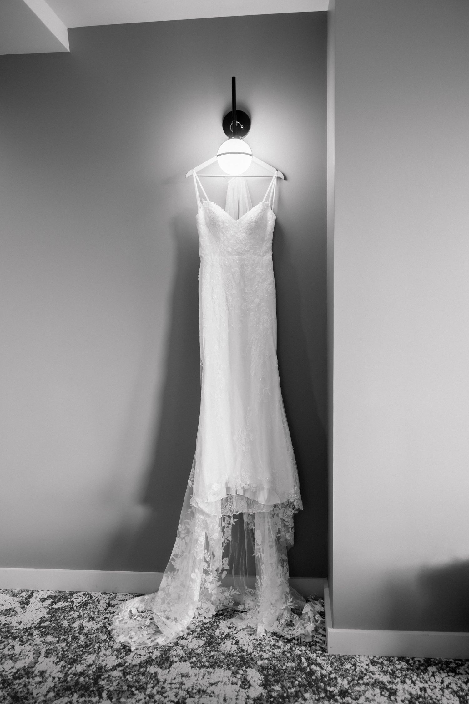 Black and white photo of an elegant lace wedding gown hanging on a wall hook beneath a lit sconce.