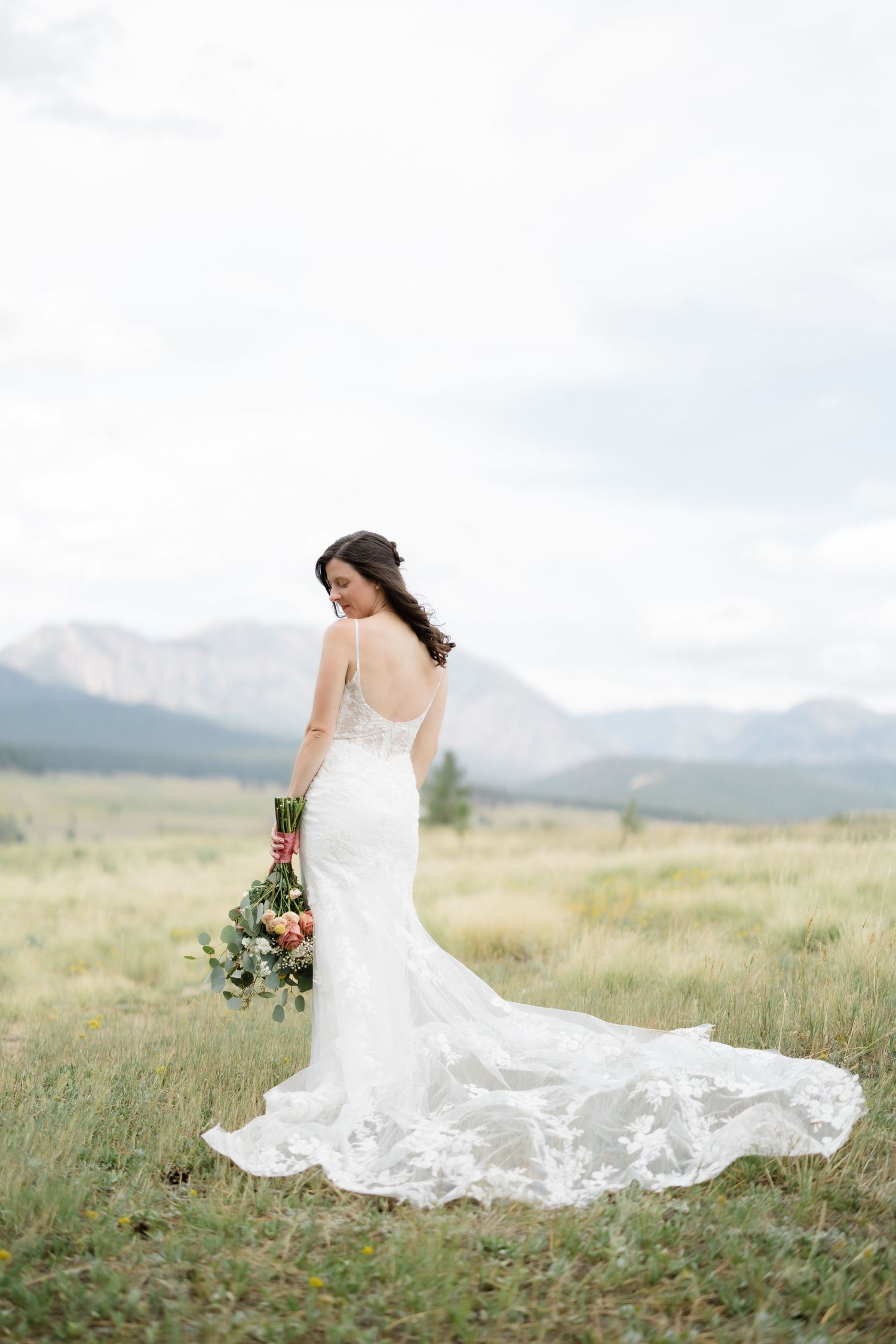 Bride in white lace mermaid gown with train holds bouquet in mountain meadow.